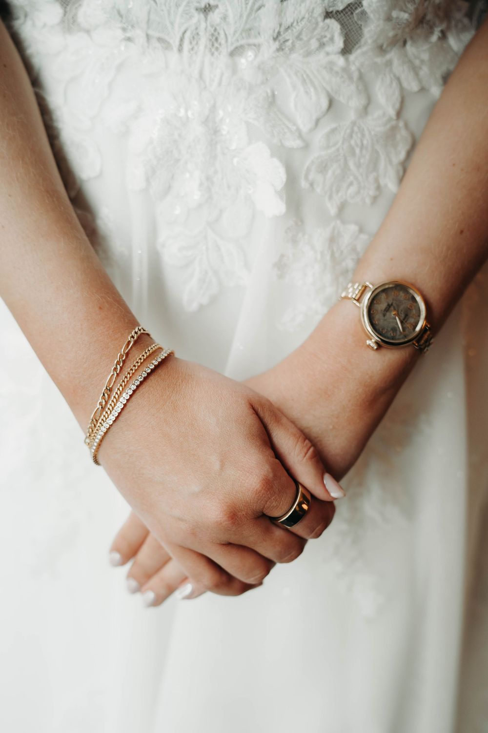 Close-up of bride's hands clasped together wearing gold watch, delicate bracelets, and ring.