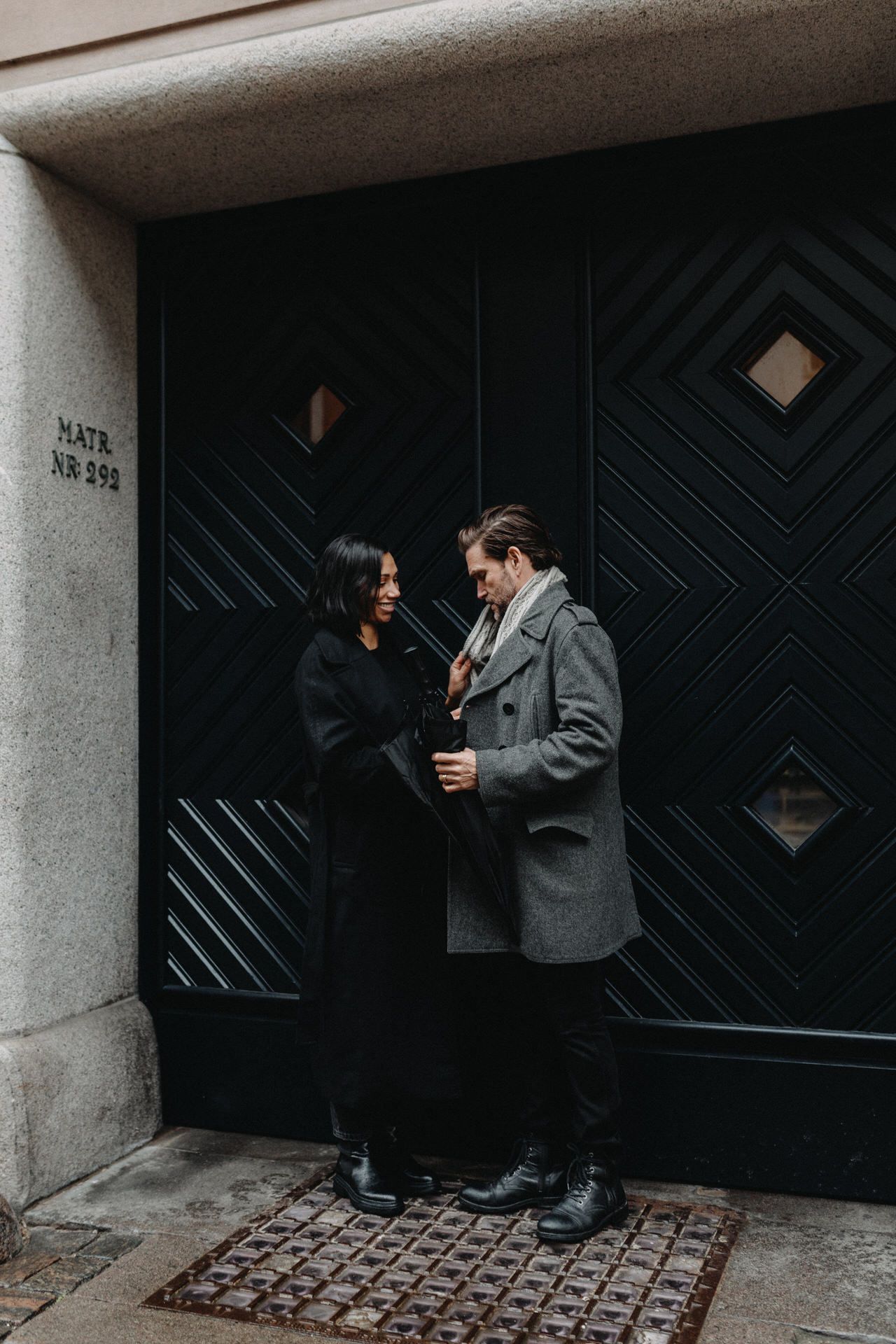 Two people share a conversation in front of a dark geometric patterned door in an urban setting.