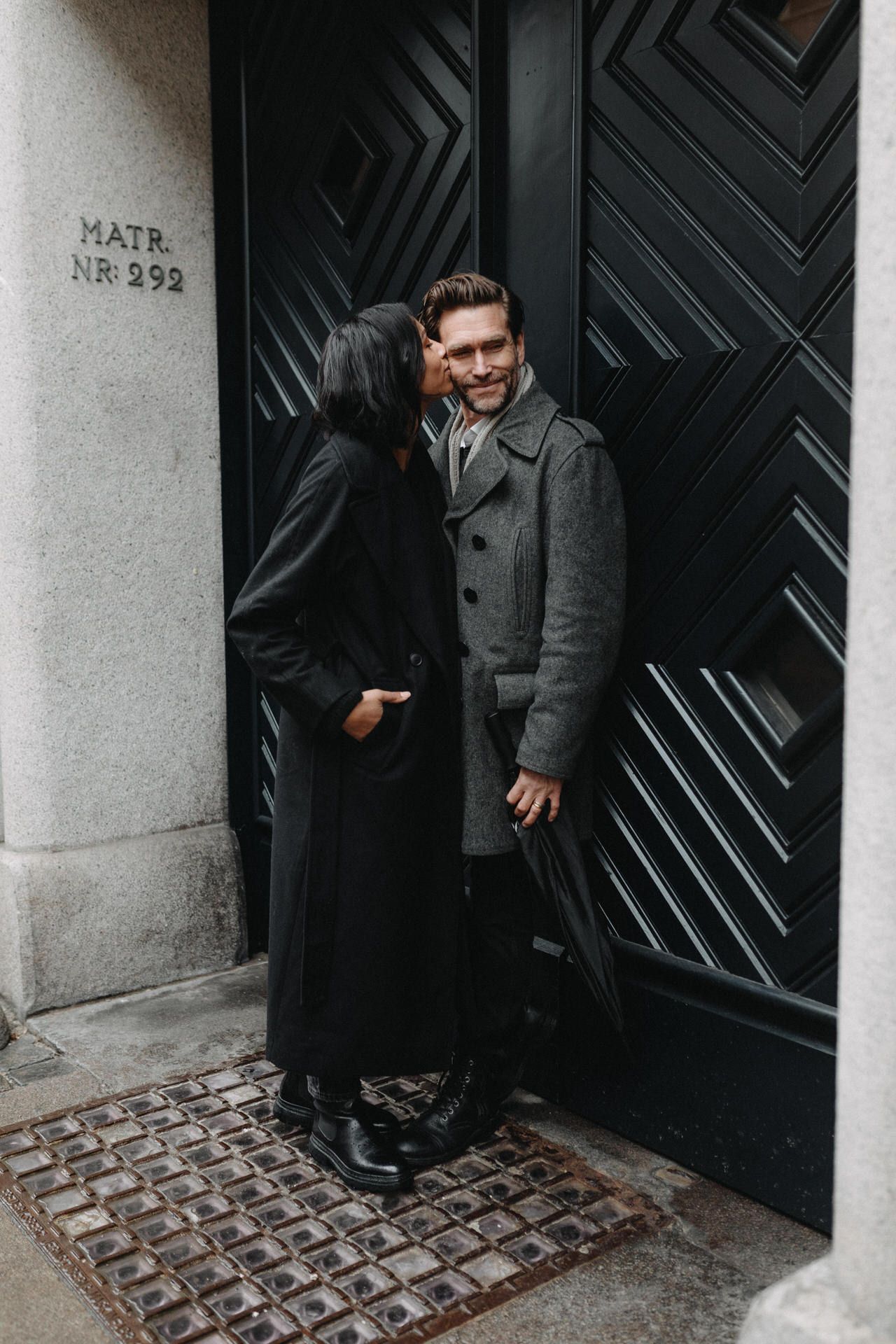 A couple shares an intimate moment in front of a geometric black door in winter coats.
