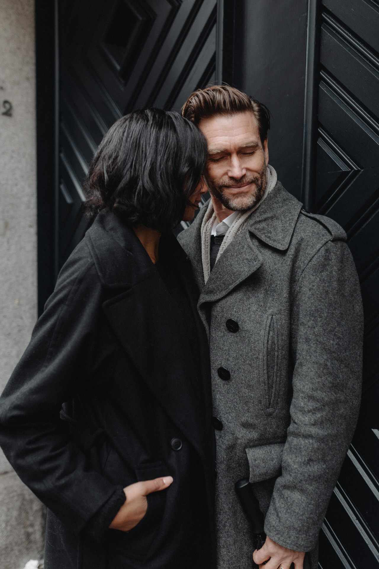 A couple in winter coats embraces and laughs together against a dark door background.