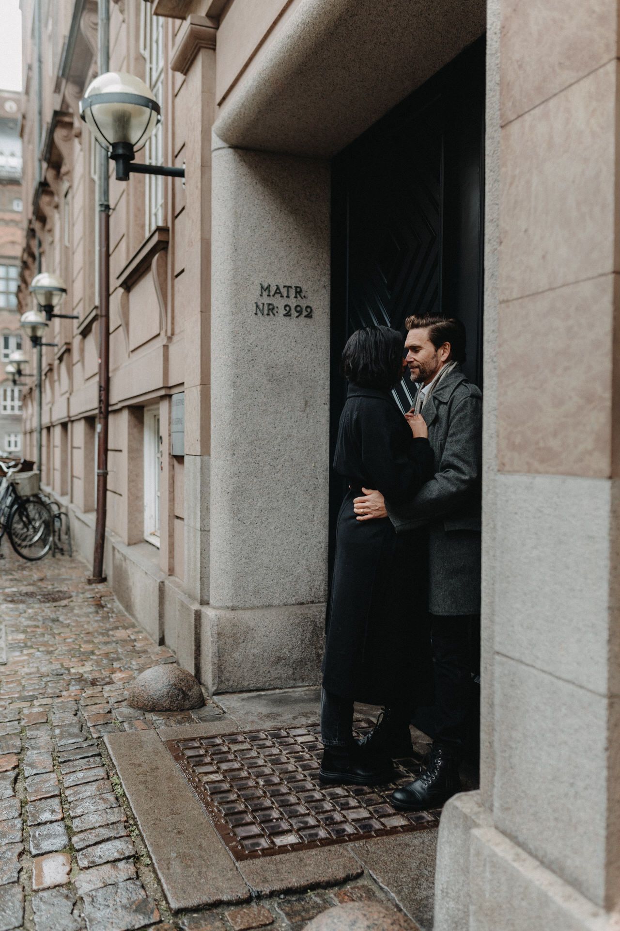 A couple embraces in a doorway of a historic European building with cobblestone streets and vintage lamp posts.