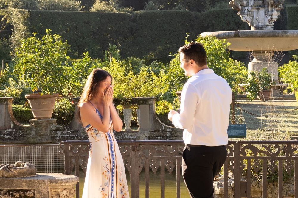 A romantic proposal moment in a sunlit garden terrace with stone balustrades and potted plants.