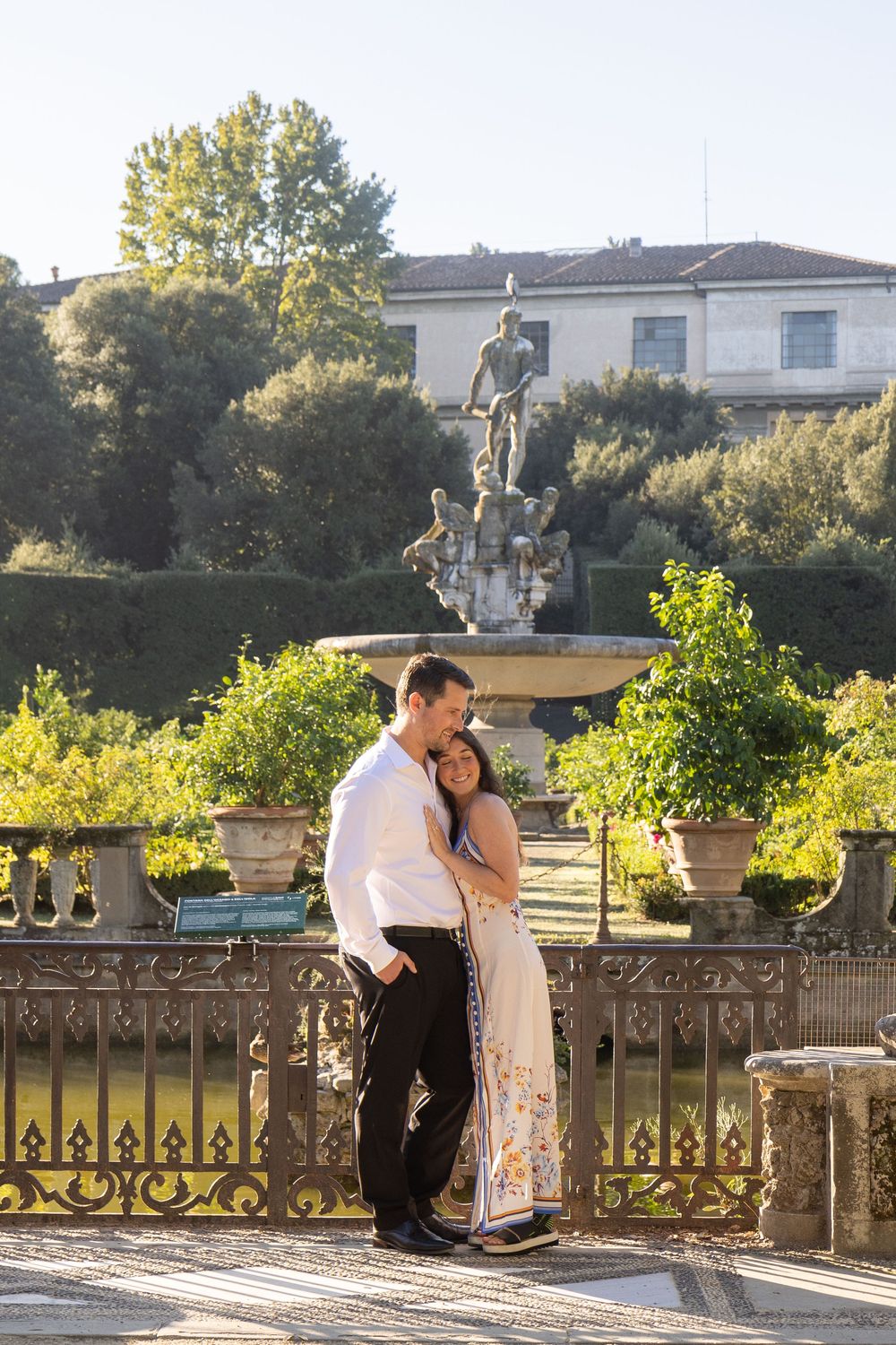 A couple embraces near an ornate fountain in a garden setting during golden hour.