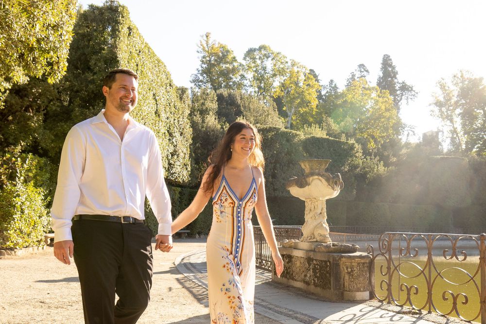 A couple walks together in a sunlit garden wearing elegant white and blue outfits during a romantic outdoor photoshoot.