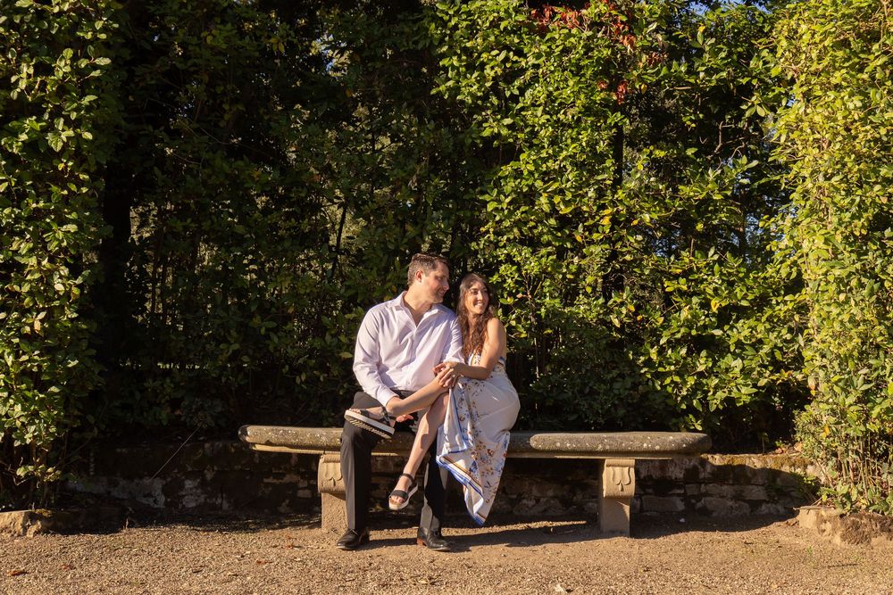 A couple sits together on a wooden bench in a sunlit garden surrounded by lush green foliage.