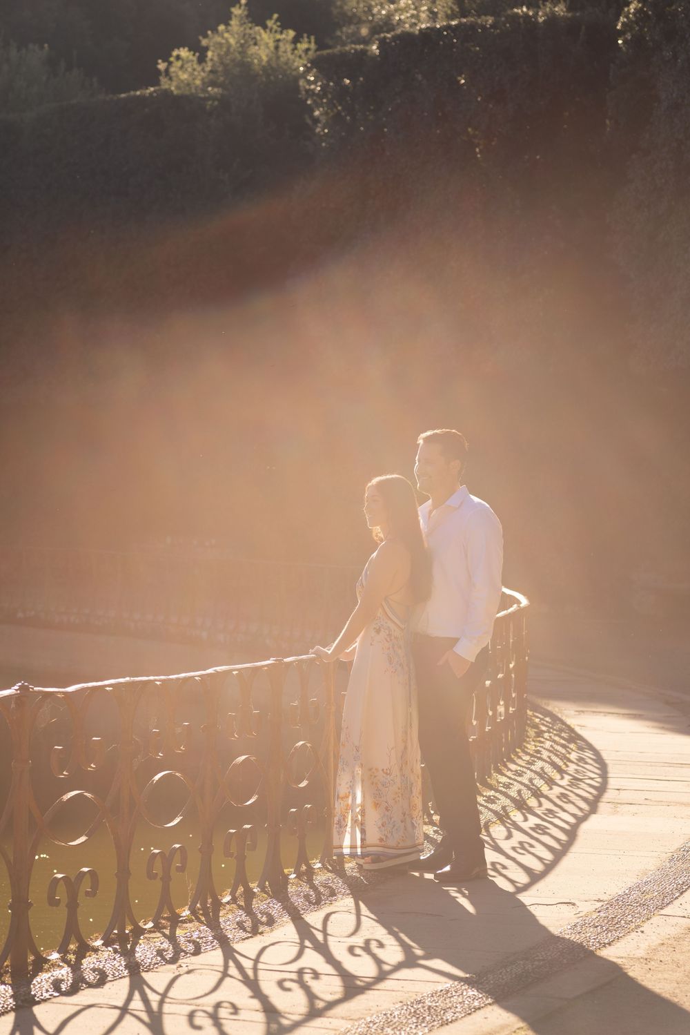 Romantic couple silhouetted against sunlight on bridge with ornate iron railing creating dramatic shadows.