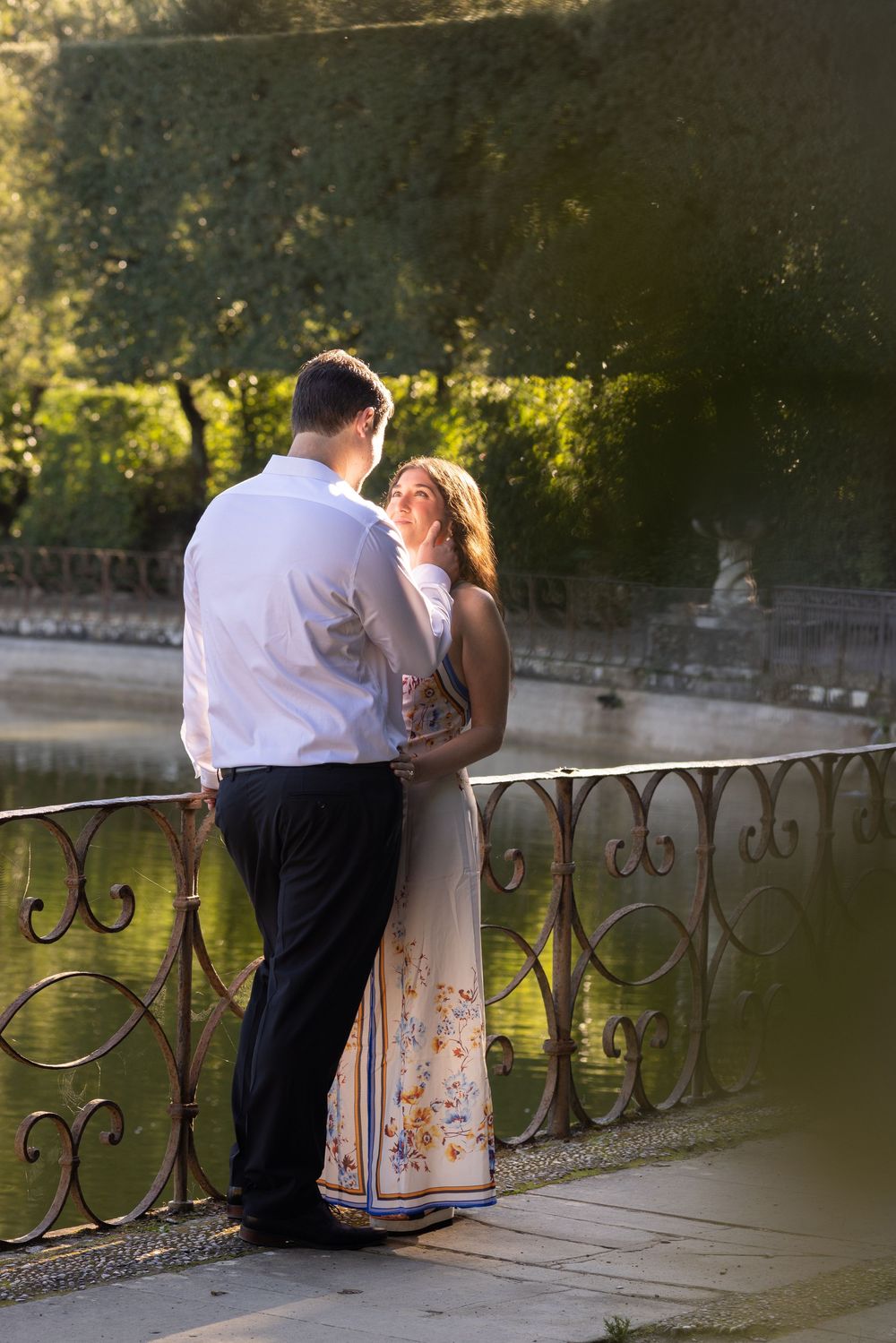 Couple in formal attire share an intimate moment along a decorative iron railing by a lake at sunset.