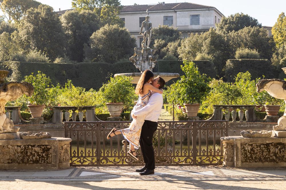 A couple shares a romantic kiss in a garden setting with stone walls and lush greenery in the background.