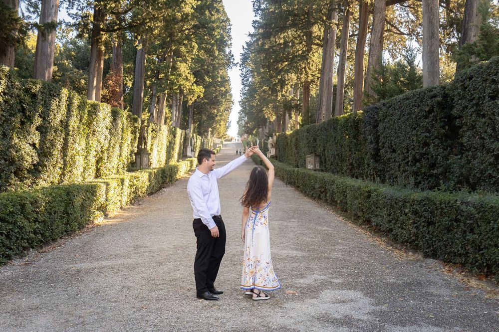 A couple walks hand in hand down a tree-lined path surrounded by lush greenery and dappled sunlight.