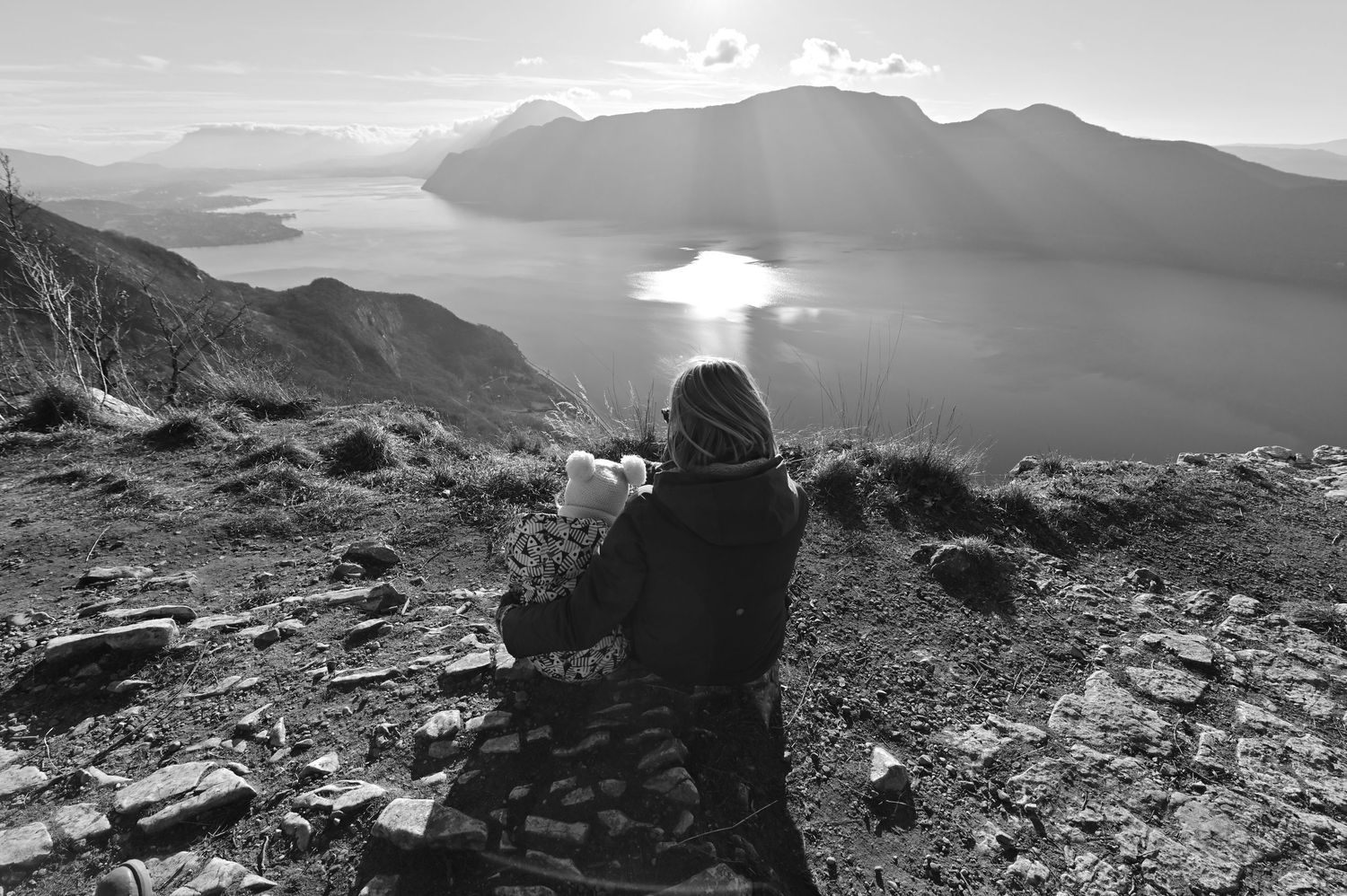 Mère assise avec son enfant face à un paysage de montagne et lac, séance photo famille en pleine nature.