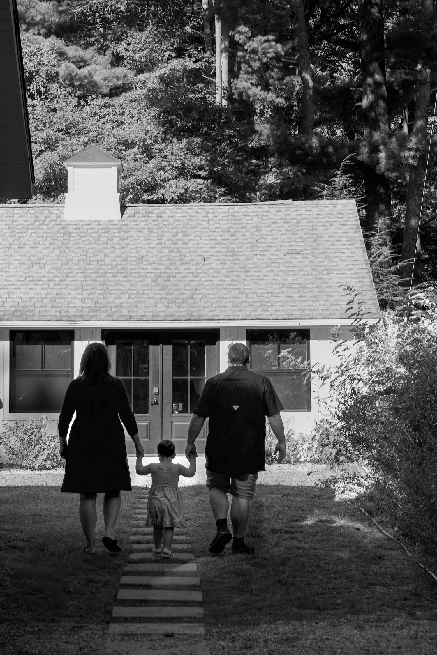 A family walks hand in hand down a garden path towards a small cottage with a shingled roof in black and white.