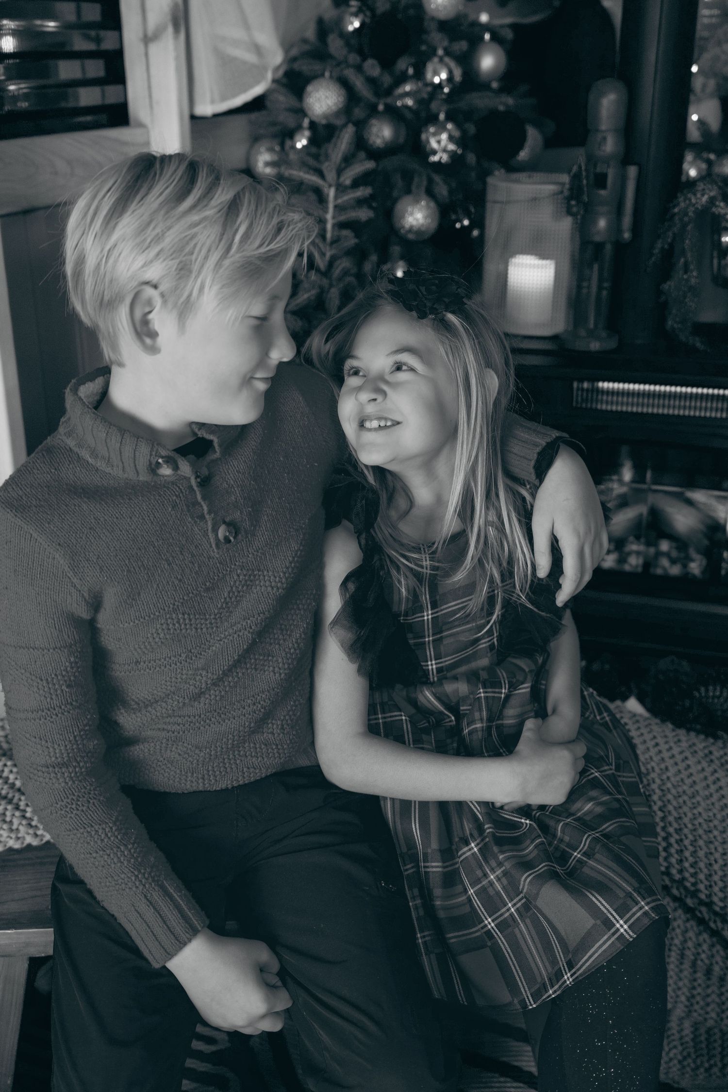 Two children share a sweet moment together in front of a decorated Christmas tree in a black and white photograph.