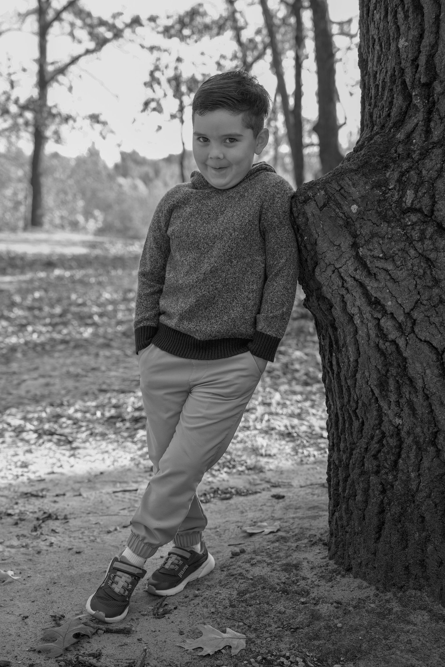 Black and white portrait of a young child in a sweater and khaki pants leaning against a tree trunk in an autumn setting.