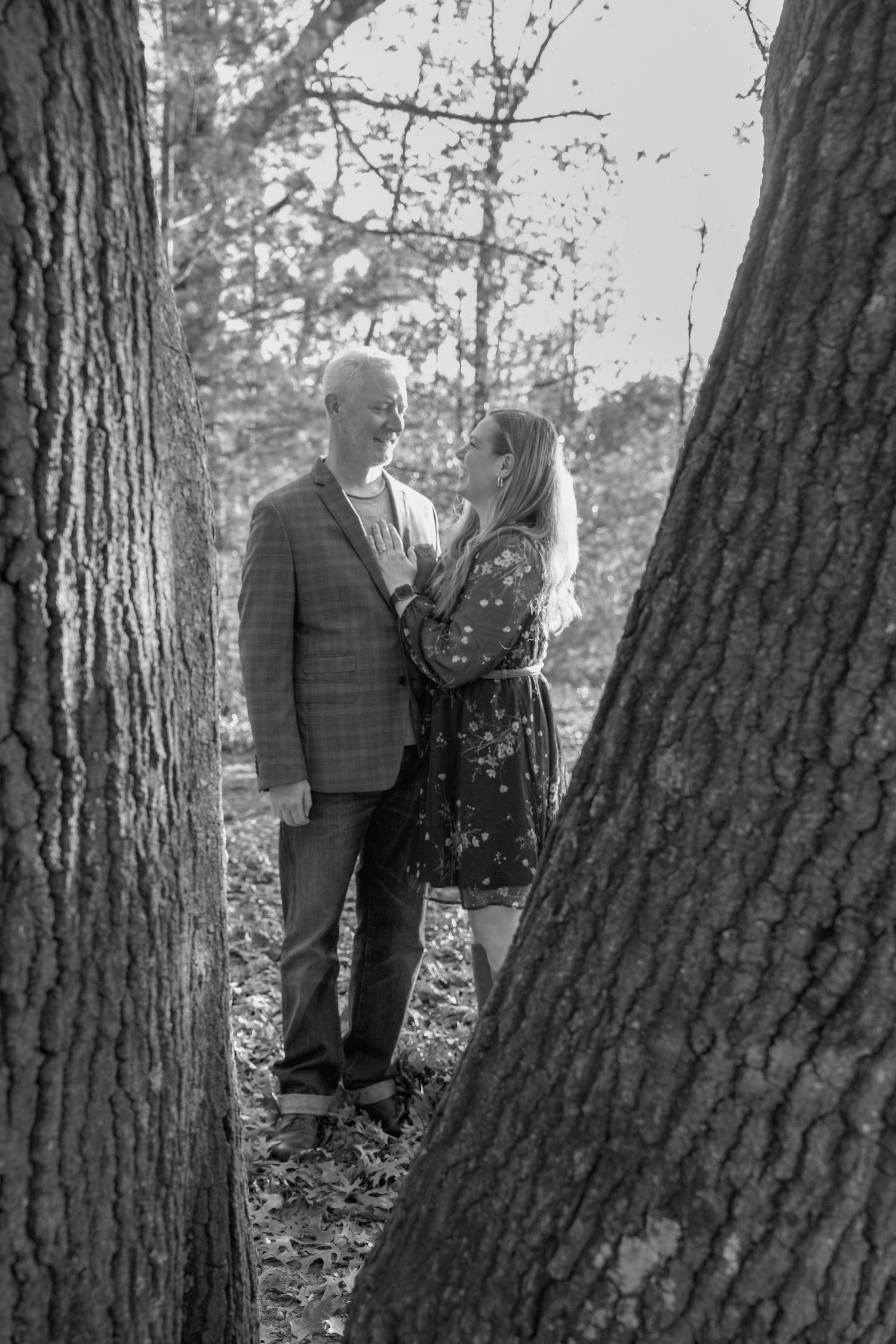 A couple stands together in a wooded area with large tree trunks creating a natural backdrop in black and white.