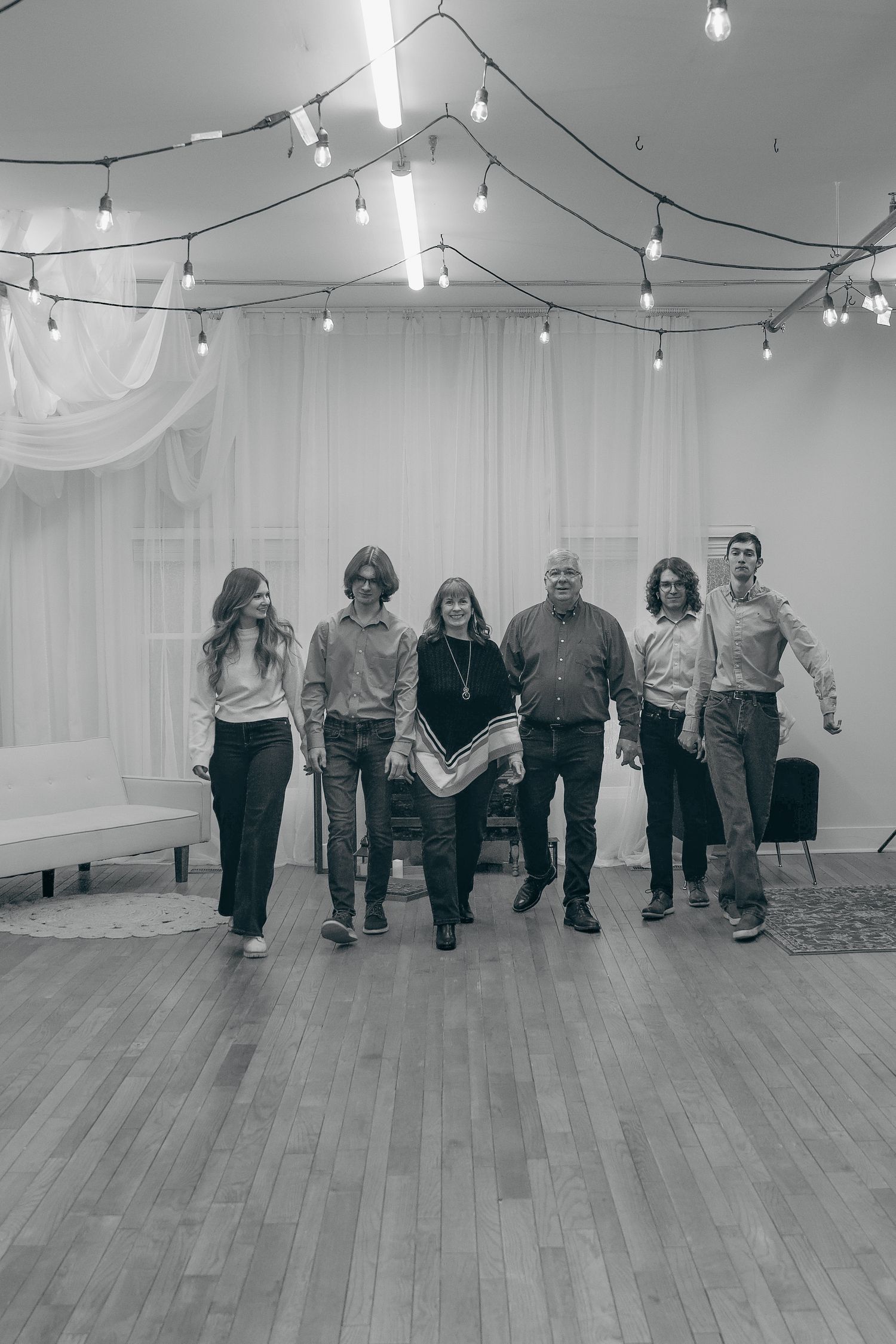 A group of people dancing in a line formation on a wooden floor under string lights in a white-walled studio space.