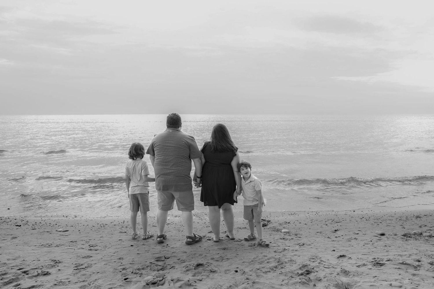 Family members sit together on a sandy beach watching the ocean waves at sunset in black and white.