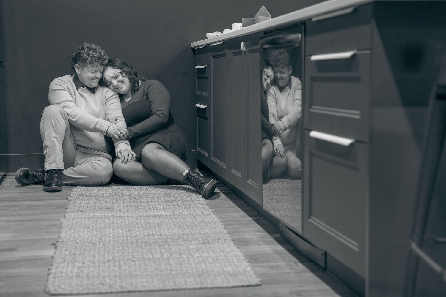 Black and white photograph showing people sitting on a kitchen floor with reflective cabinets.