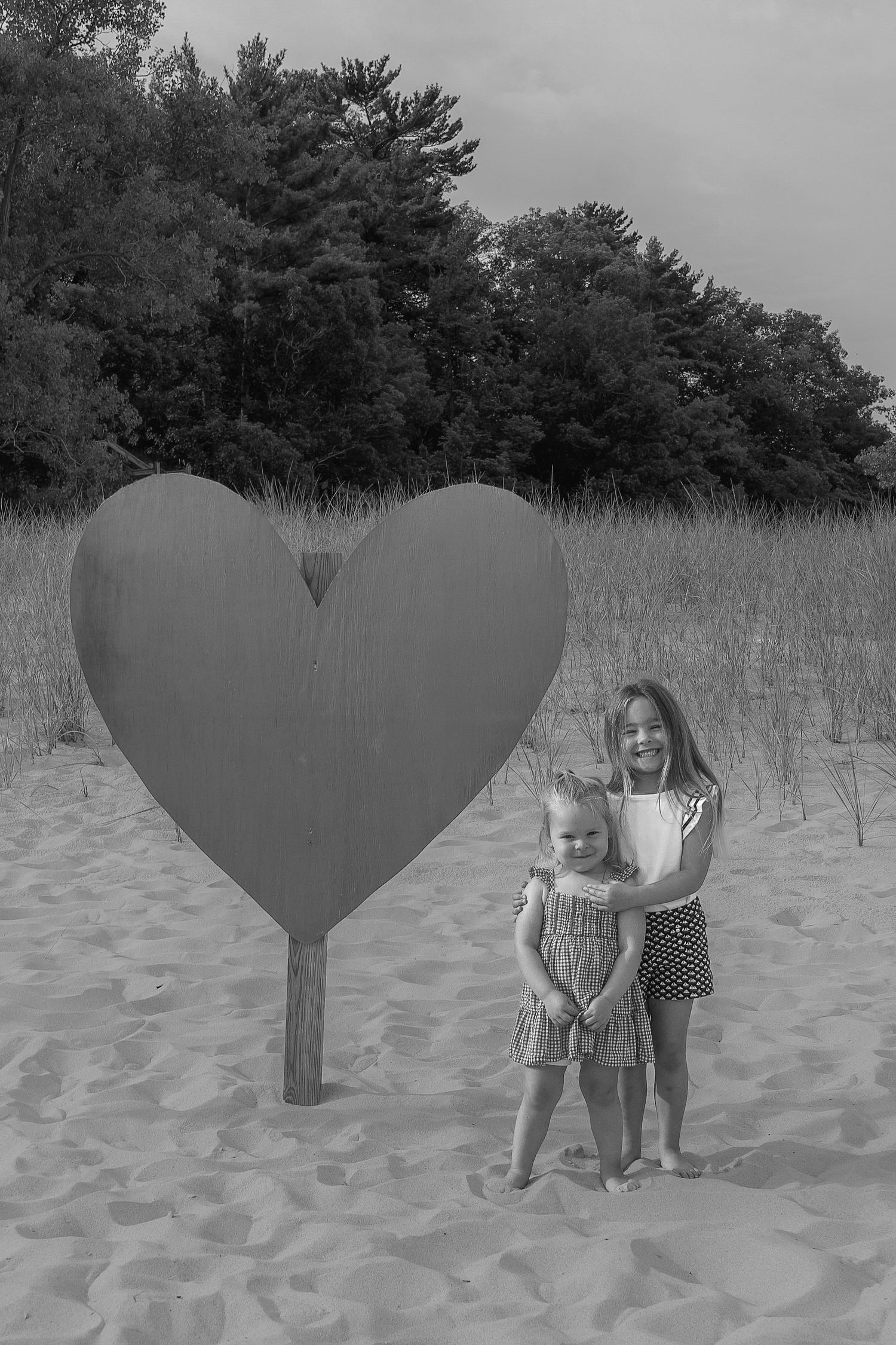 A sequence of black and white beach photos shows children posing behind a large heart-shaped cutout in the sand.