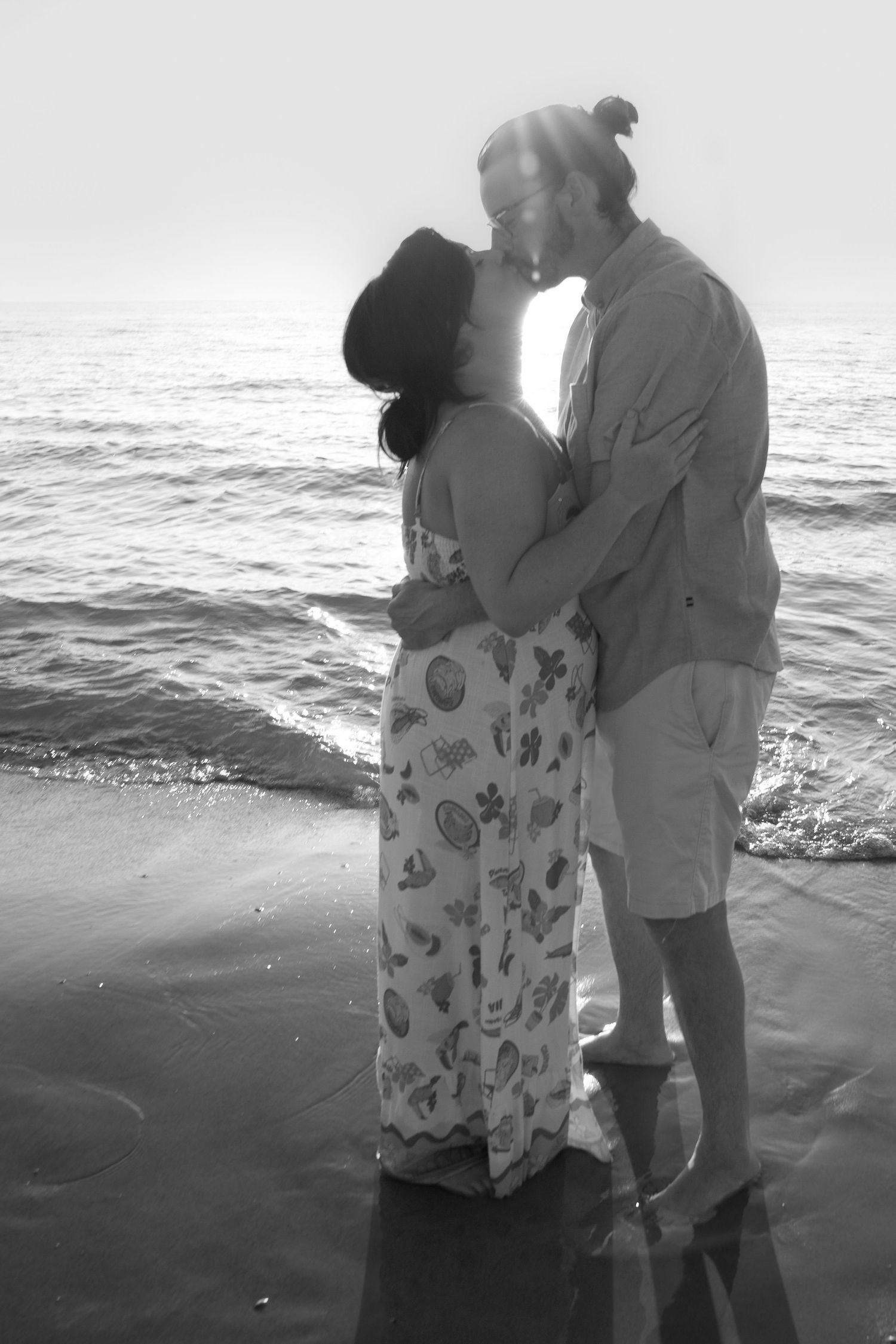 A romantic couple shares a tender embrace on a beach at sunset in black and white photography.
