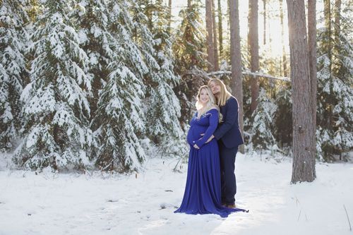 A couple in formal attire embraces in a snowy winter forest with snow-covered evergreen trees in the background.