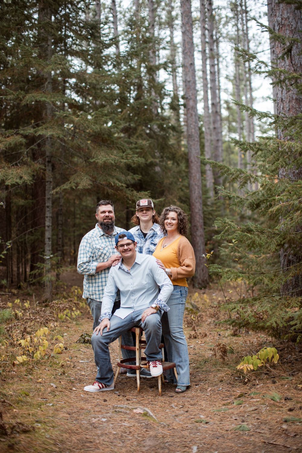 A family poses together on a wooden chair in a pine forest setting during an outdoor photo session.