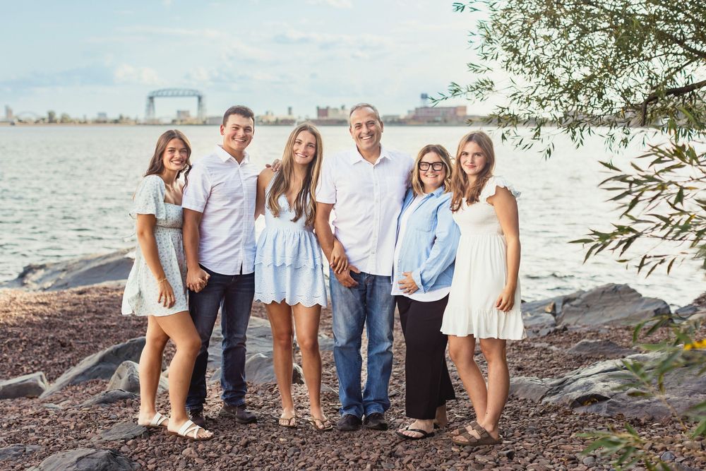 A family in white and blue outfits poses together on rocky shoreline with water and trees in background.