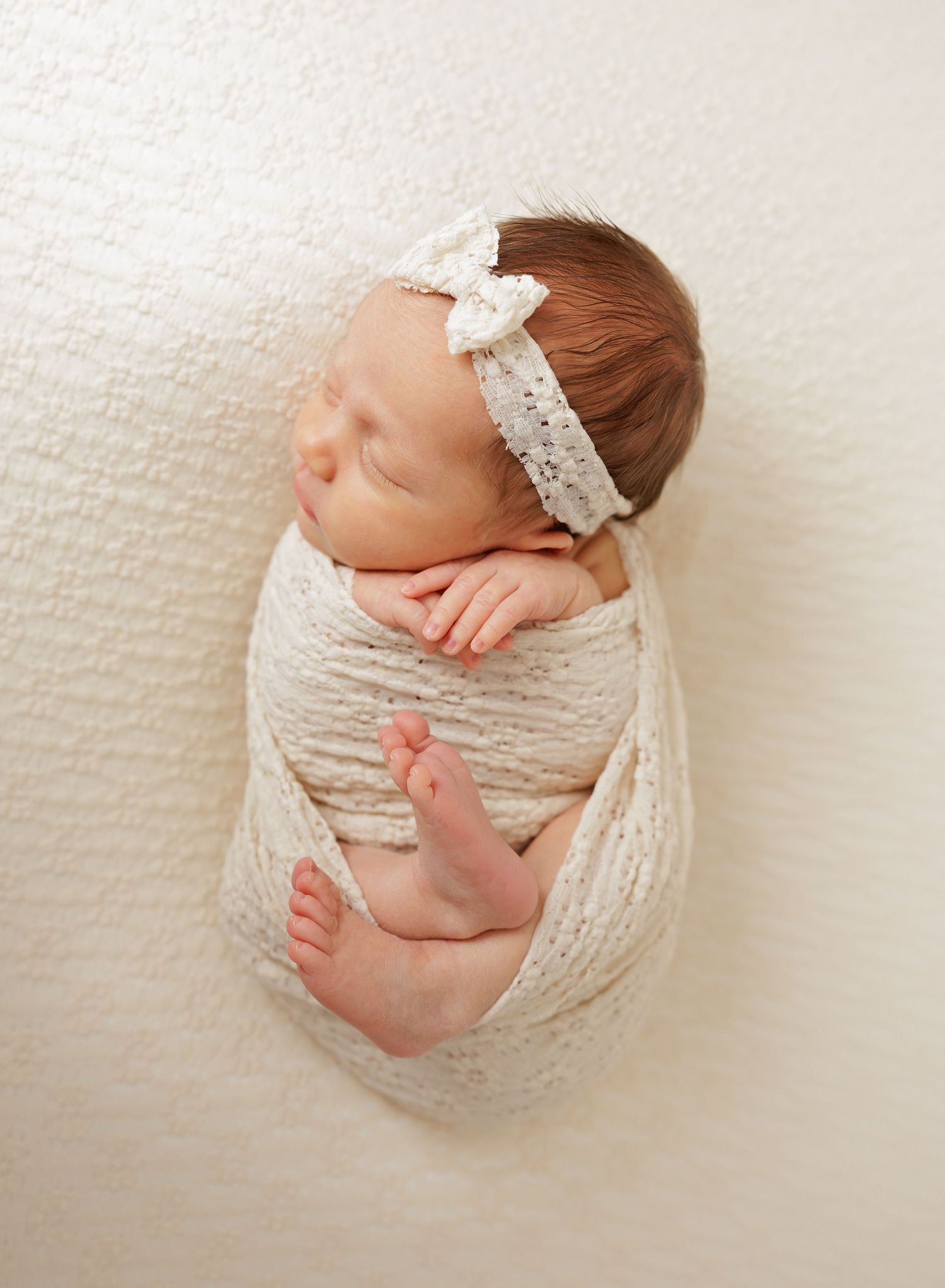 Newborn baby wrapped in cream colored fabric with lace headband on white background.