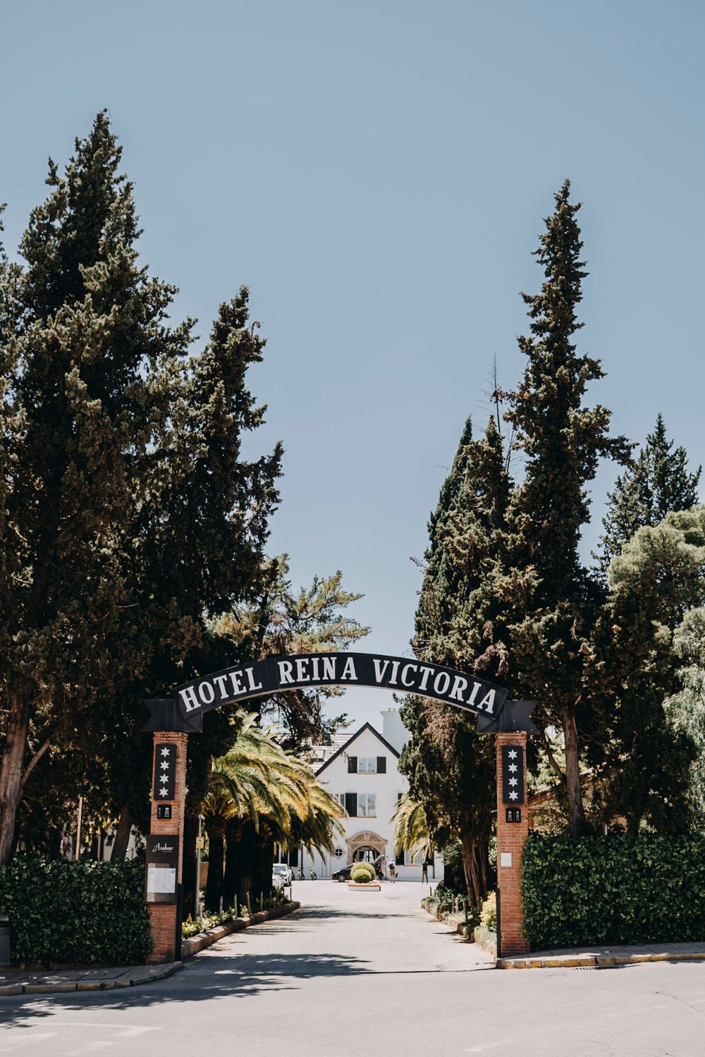 Entrance gate to Hotel Reina Victoria with palm trees and cypress trees lining the driveway.