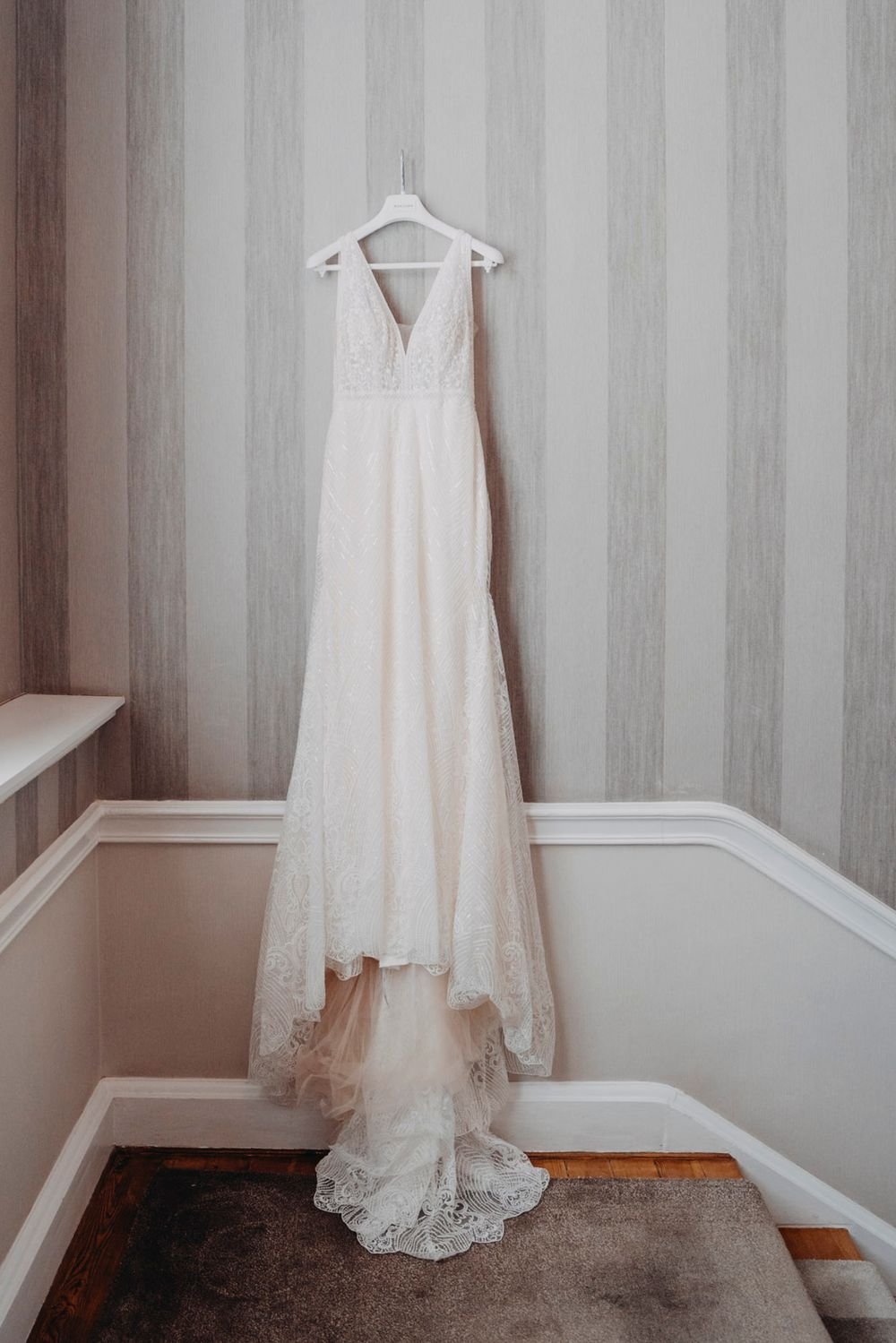 White lace wedding dress hanging against striped gray wallpaper in preparation room.