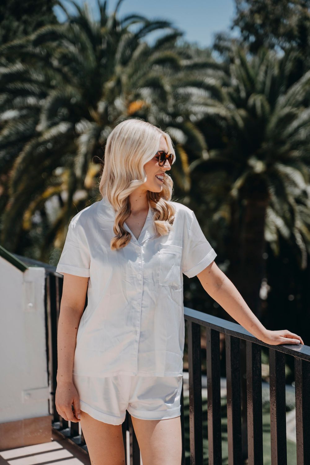 A casual summer outfit featuring white romper against a tropical palm tree backdrop on a sunny balcony.