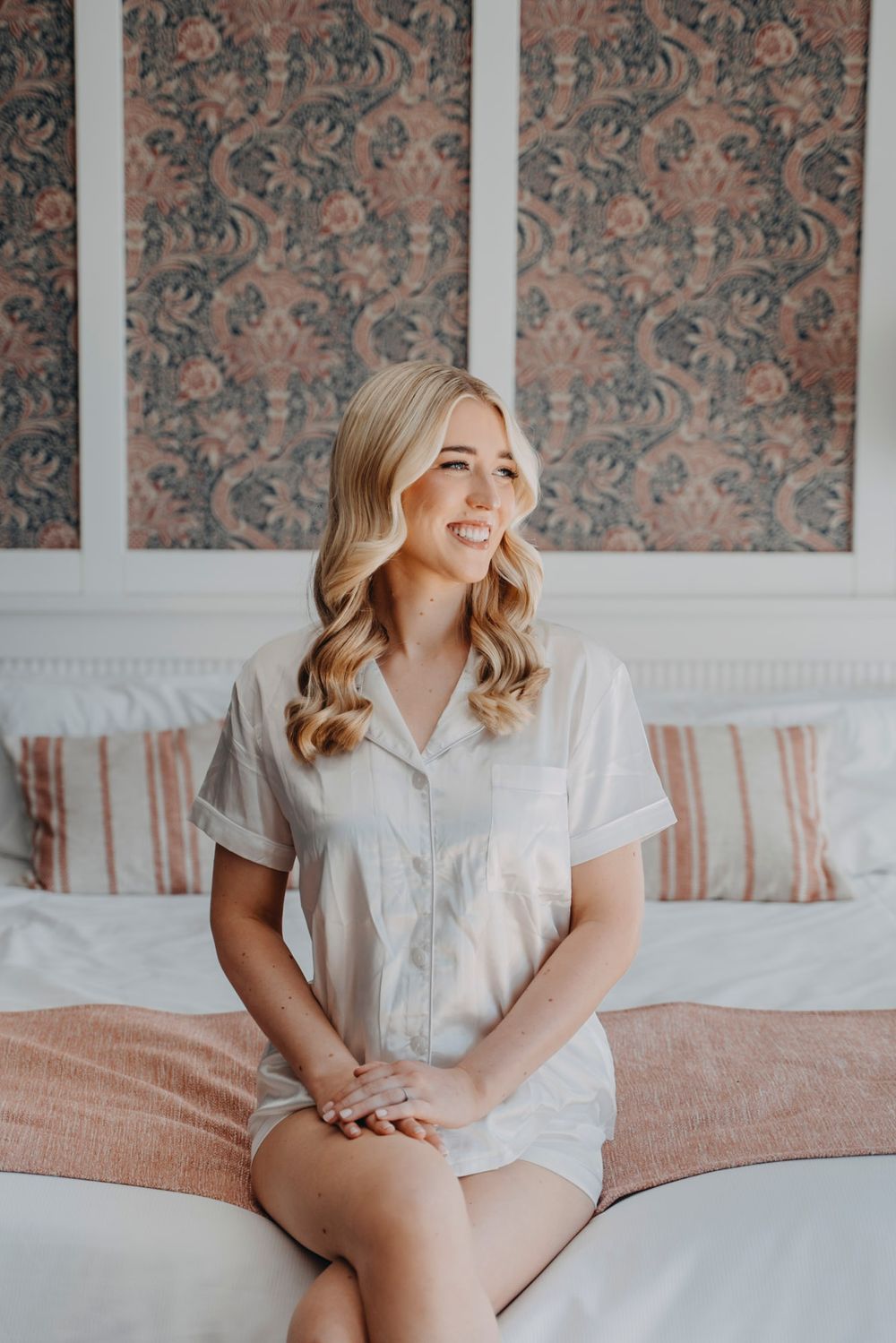 An indoor portrait showing a white pajama set against floral wallpaper and pink bedding.