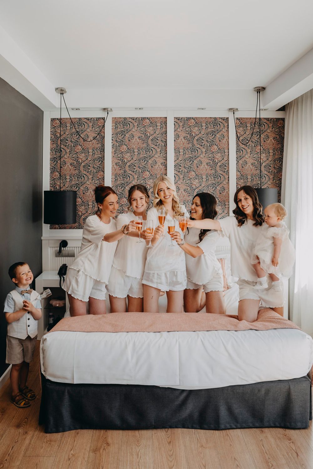 A group wearing matching white outfits poses playfully together on a large bed during pre-wedding preparations.