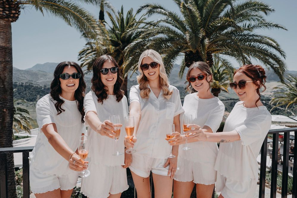 Five friends in matching white outfits enjoying drinks on a tropical balcony with palm trees and mountain views.