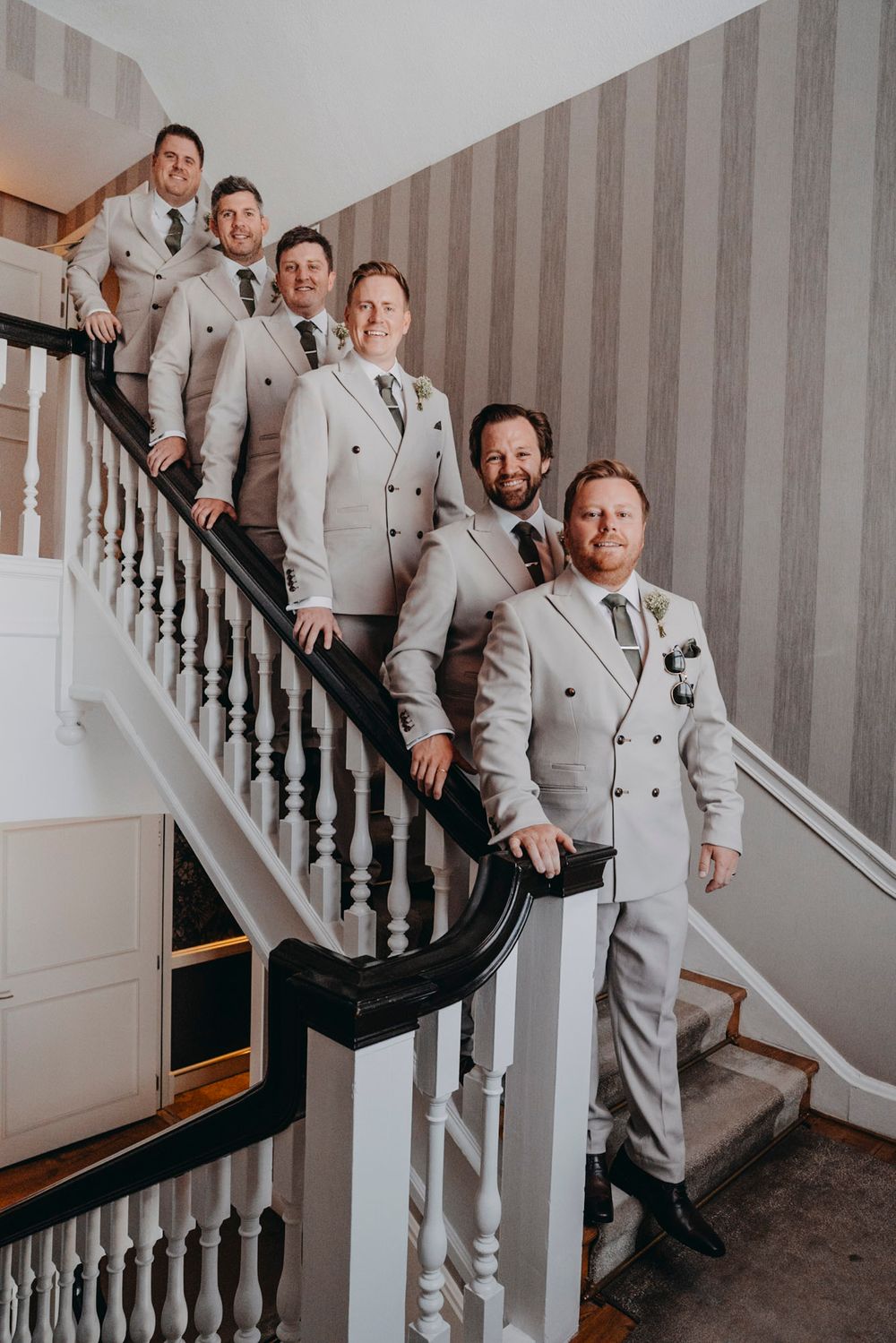 Groomsmen in light gray suits pose together on an elegant staircase with black railing.
