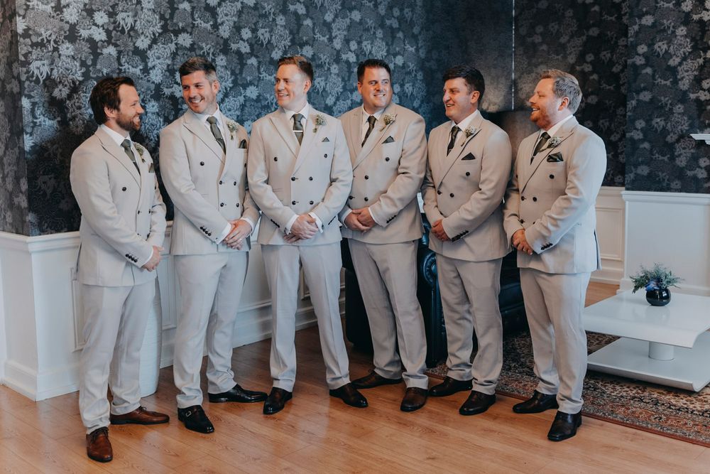 Groomsmen in light gray suits pose together against stone wall backdrop.