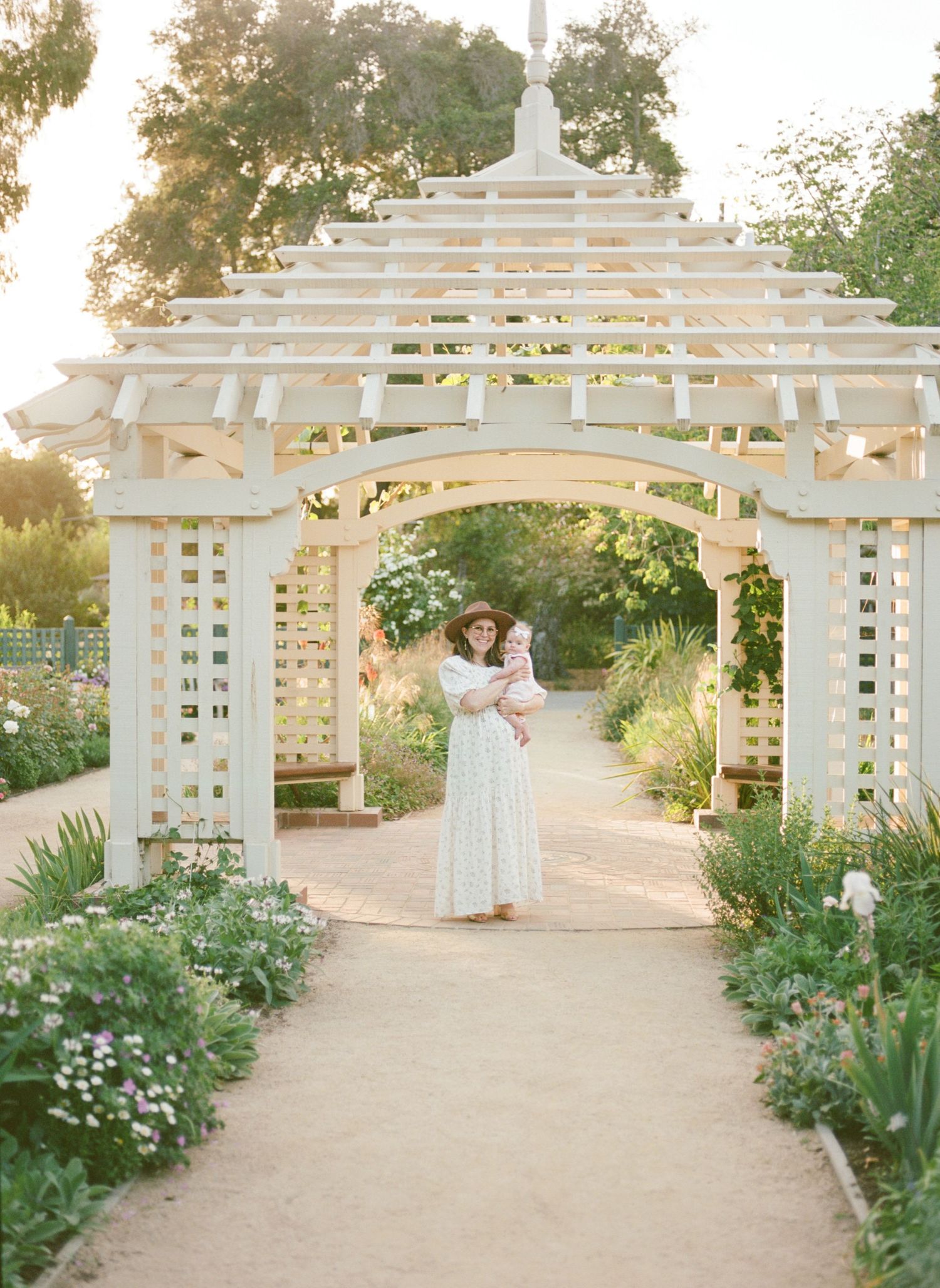 A garden scene with a white pergola archway surrounded by blooming flowers at sunset with a mother and baby captured by Chelsee Rawe