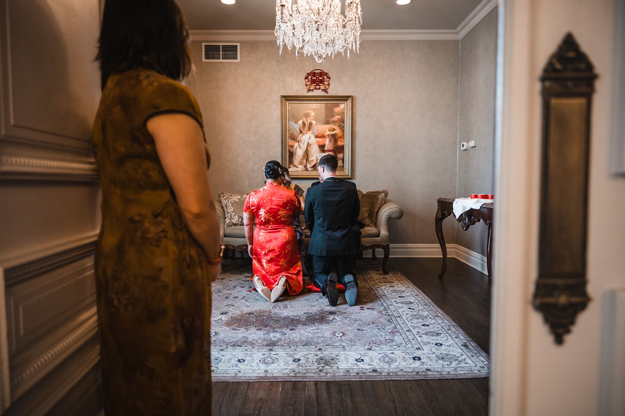 Traditional tea ceremony with guests kneeling on ornate carpet under crystal chandelier.