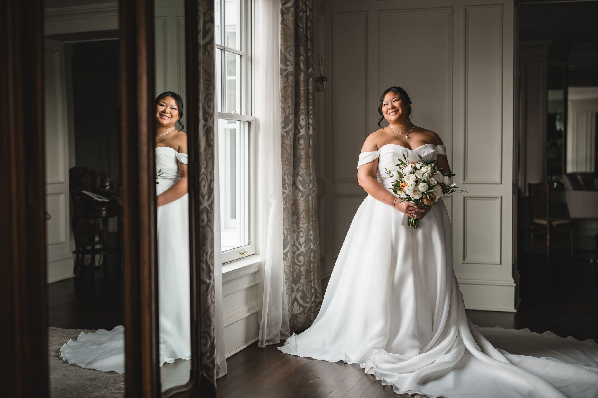 Two elegant off-shoulder wedding dresses are showcased in a classic interior with ornate moldings and natural window light.