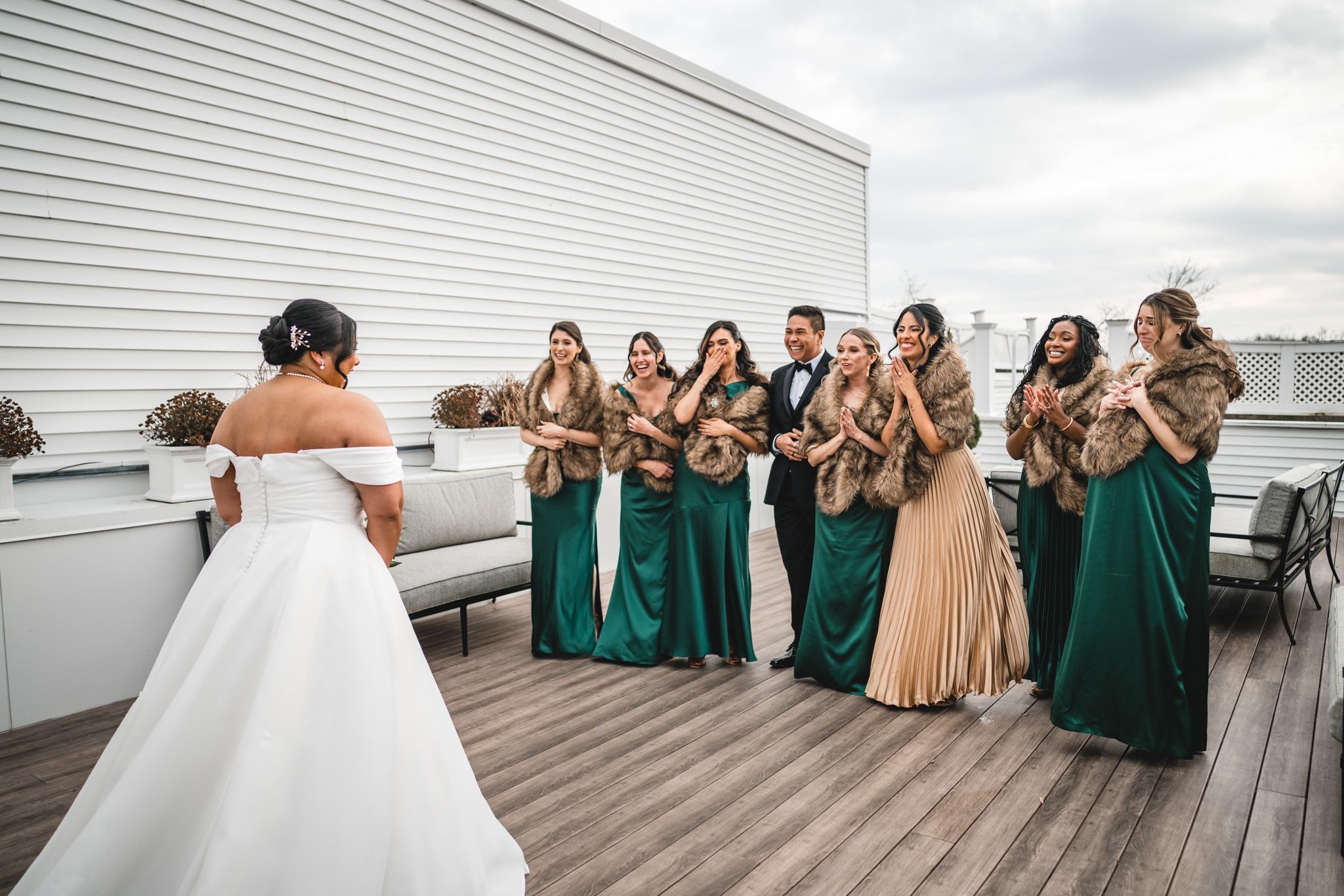 Bridesmaids in emerald green and gold dresses react emotionally on a rooftop deck.