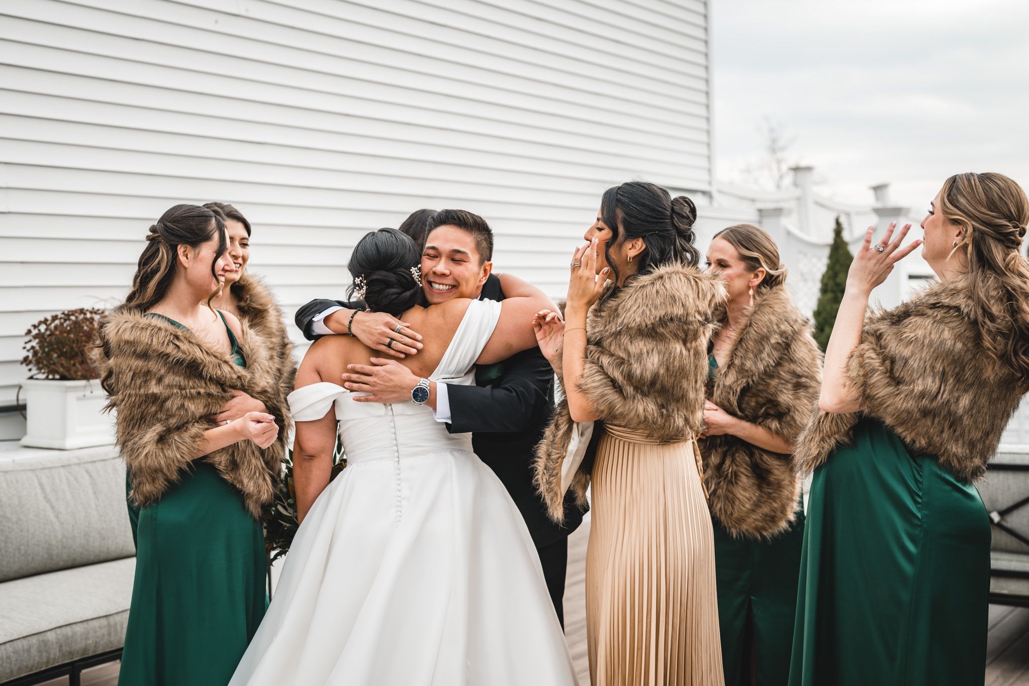 Wedding party wearing fur shawls and formal attire share an emotional group moment on an outdoor deck.