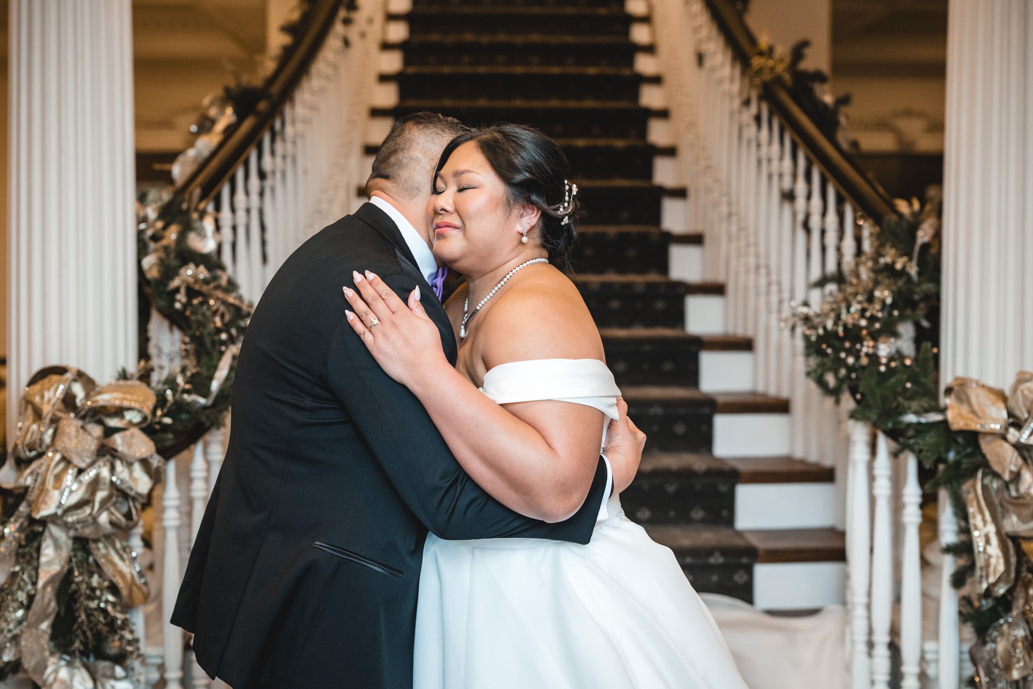 A tender embrace shared at the base of an elegantly decorated staircase during a wedding celebration.