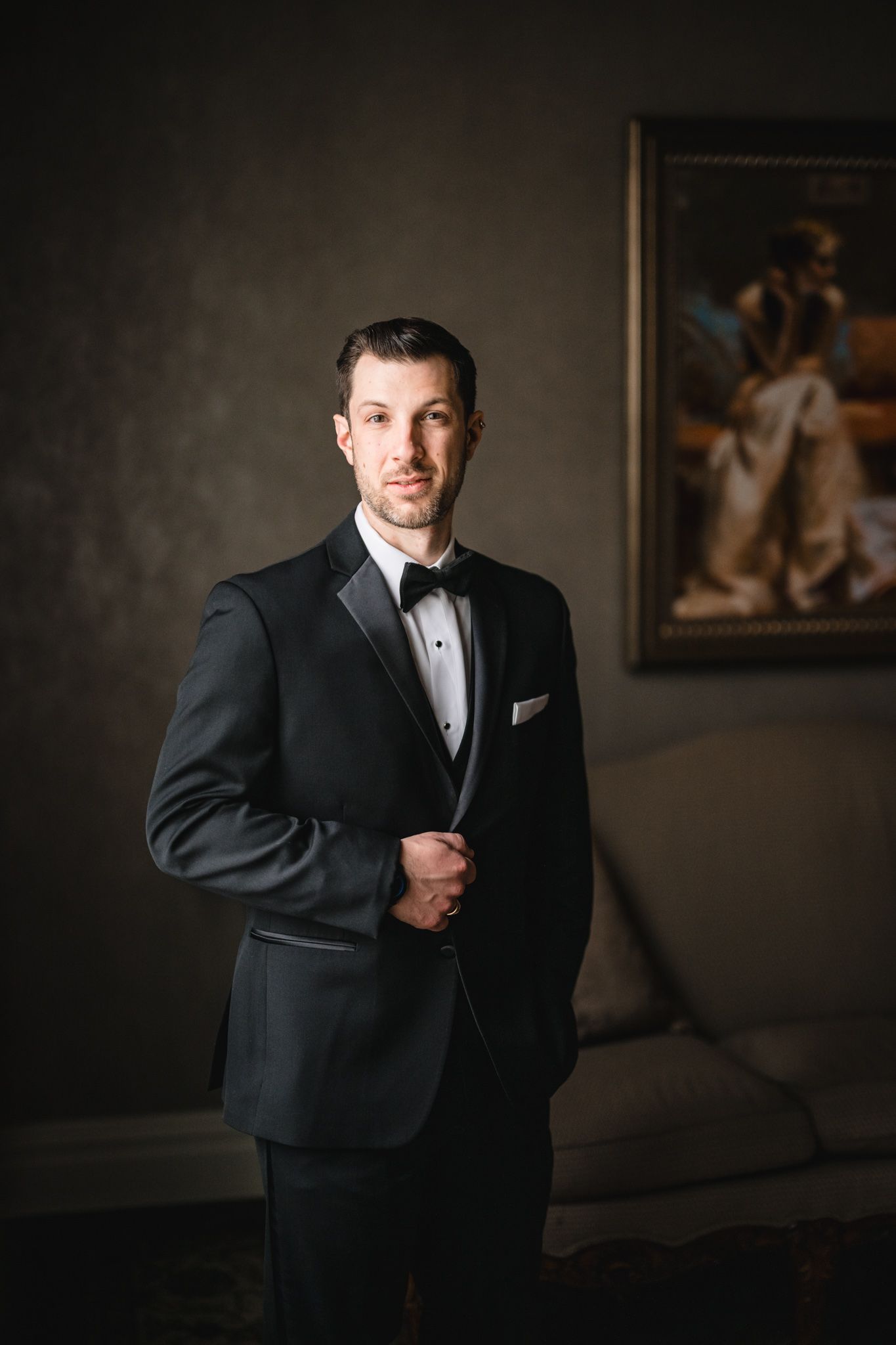 Formal portrait of someone in a black tuxedo standing against a dark wall with artwork.