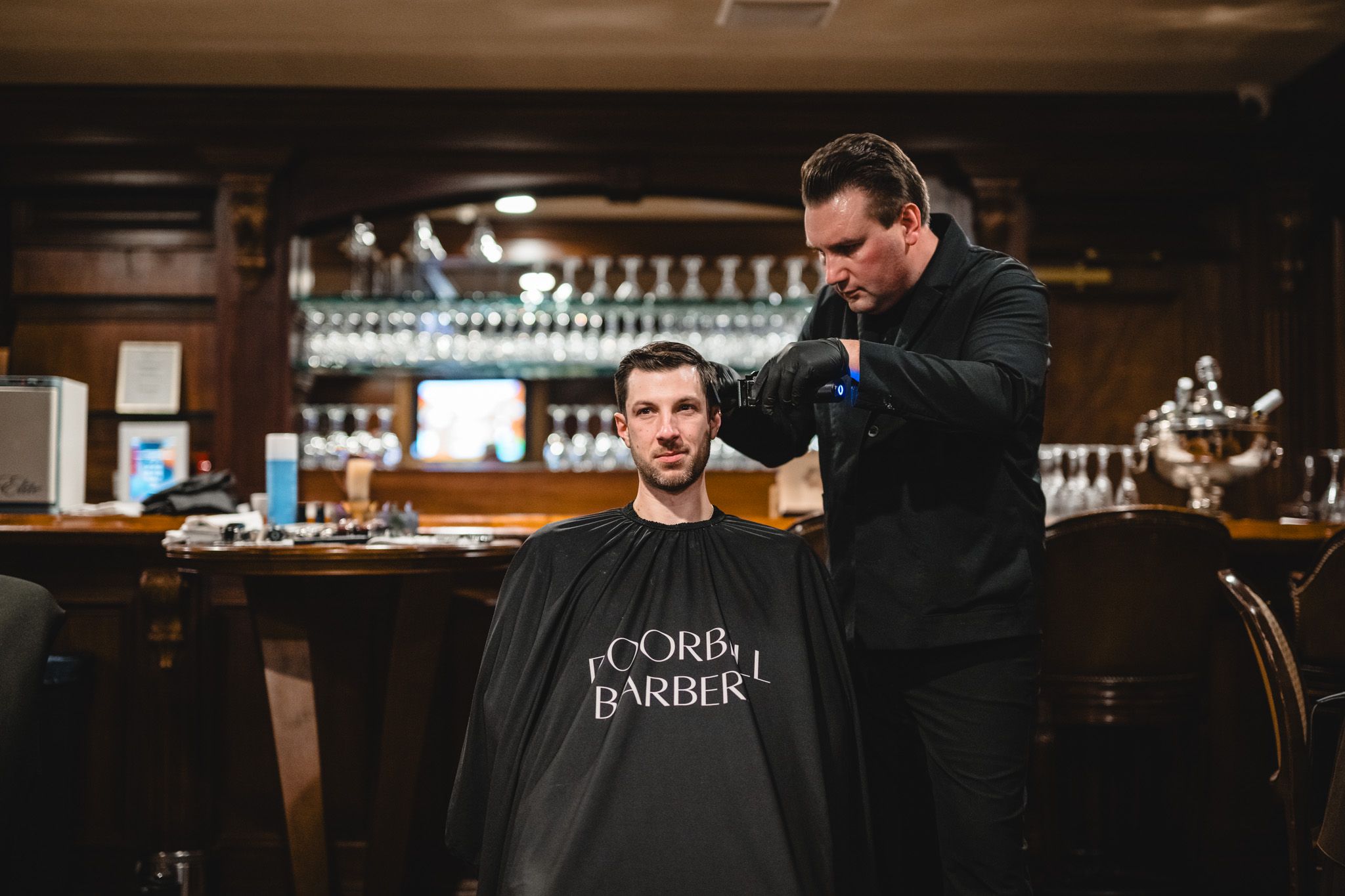A barbershop scene showing a haircut in progress at a traditional wooden bar counter.
