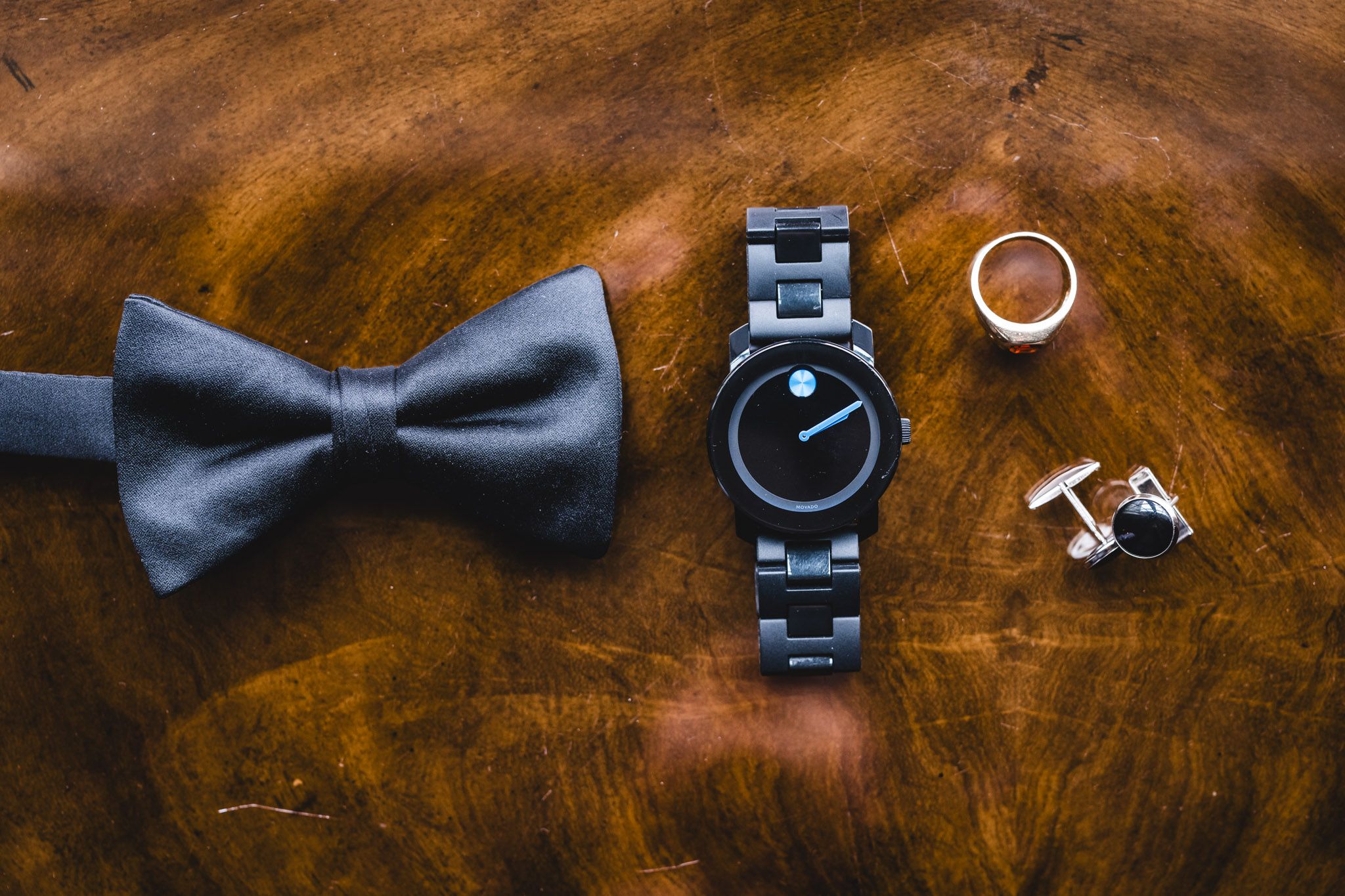 Groom's accessories including black bow tie, watch, and cufflinks arranged on wooden surface.