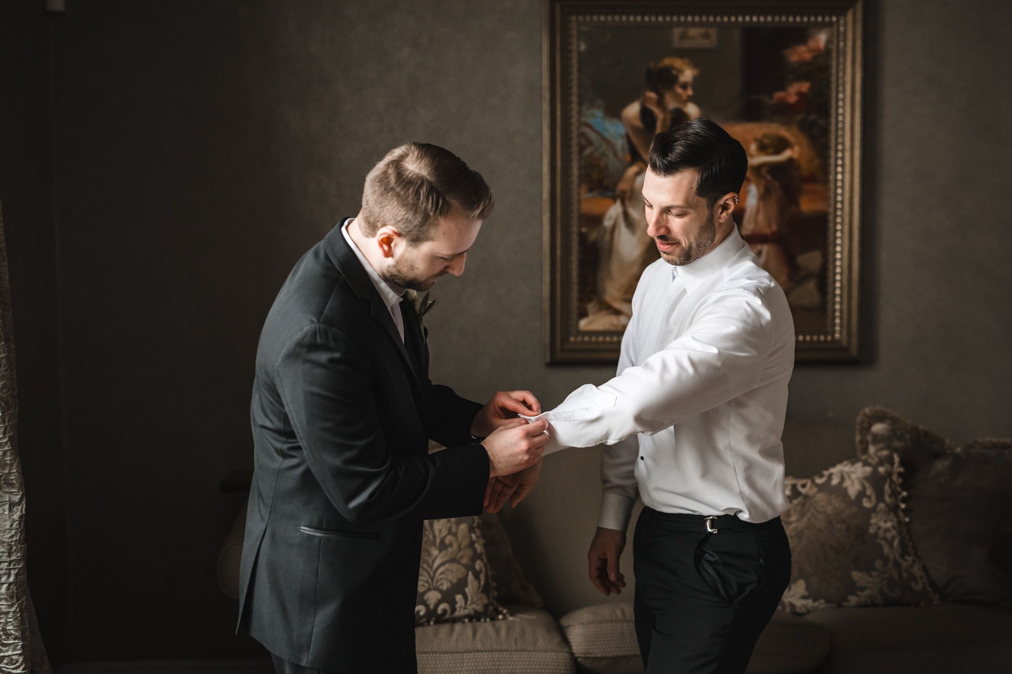Two people adjusting formal attire and helping with cufflinks in an elegant room.