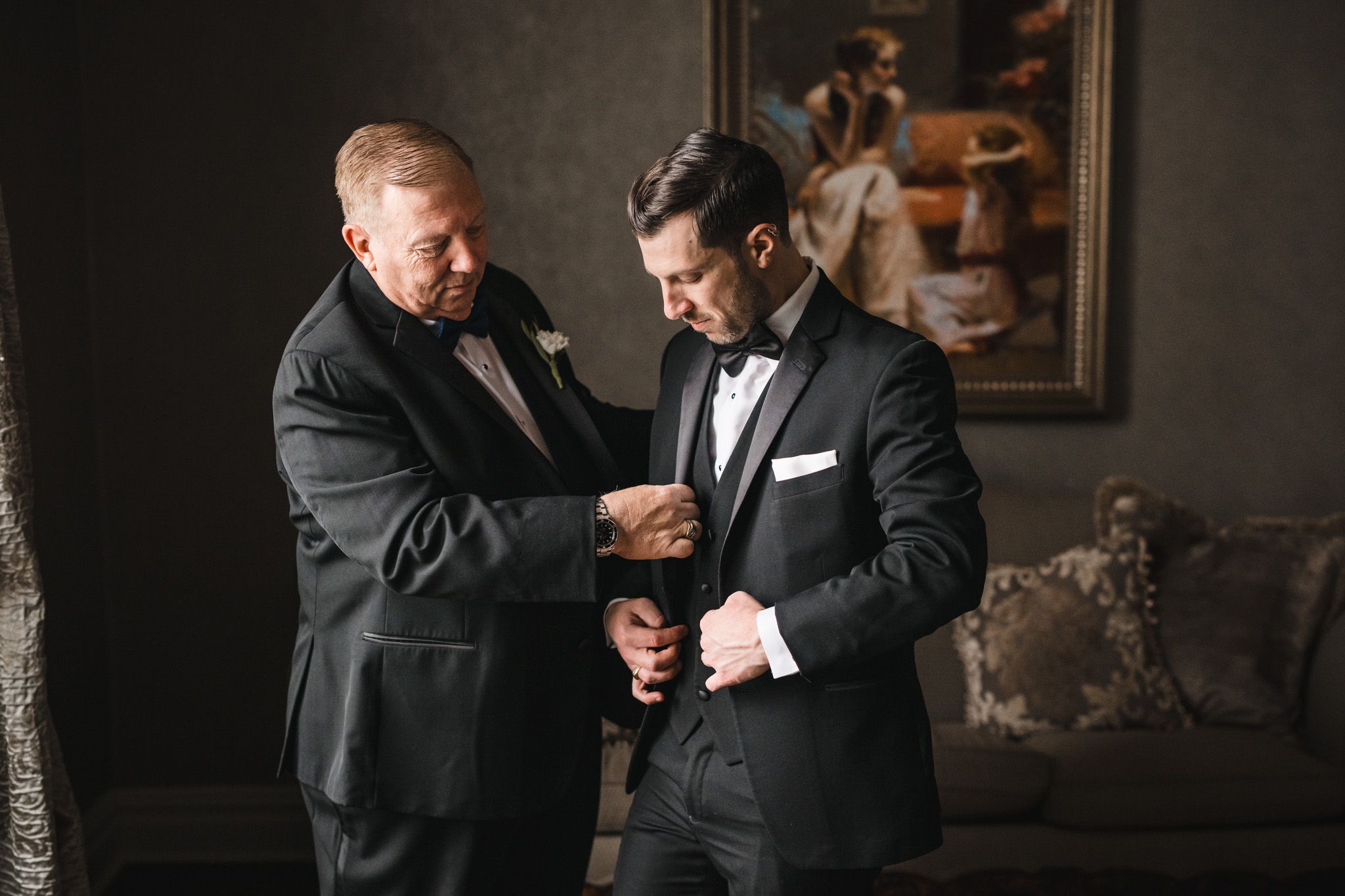 Two people in tuxedos helping each other with formal wear adjustments.