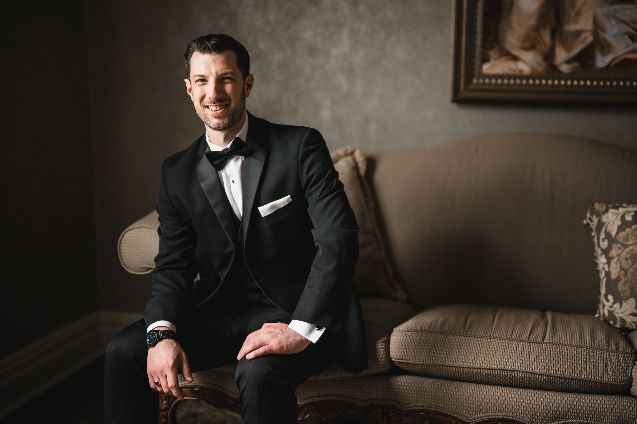 Elegant formal portrait of someone seated on a beige couch in a tuxedo.