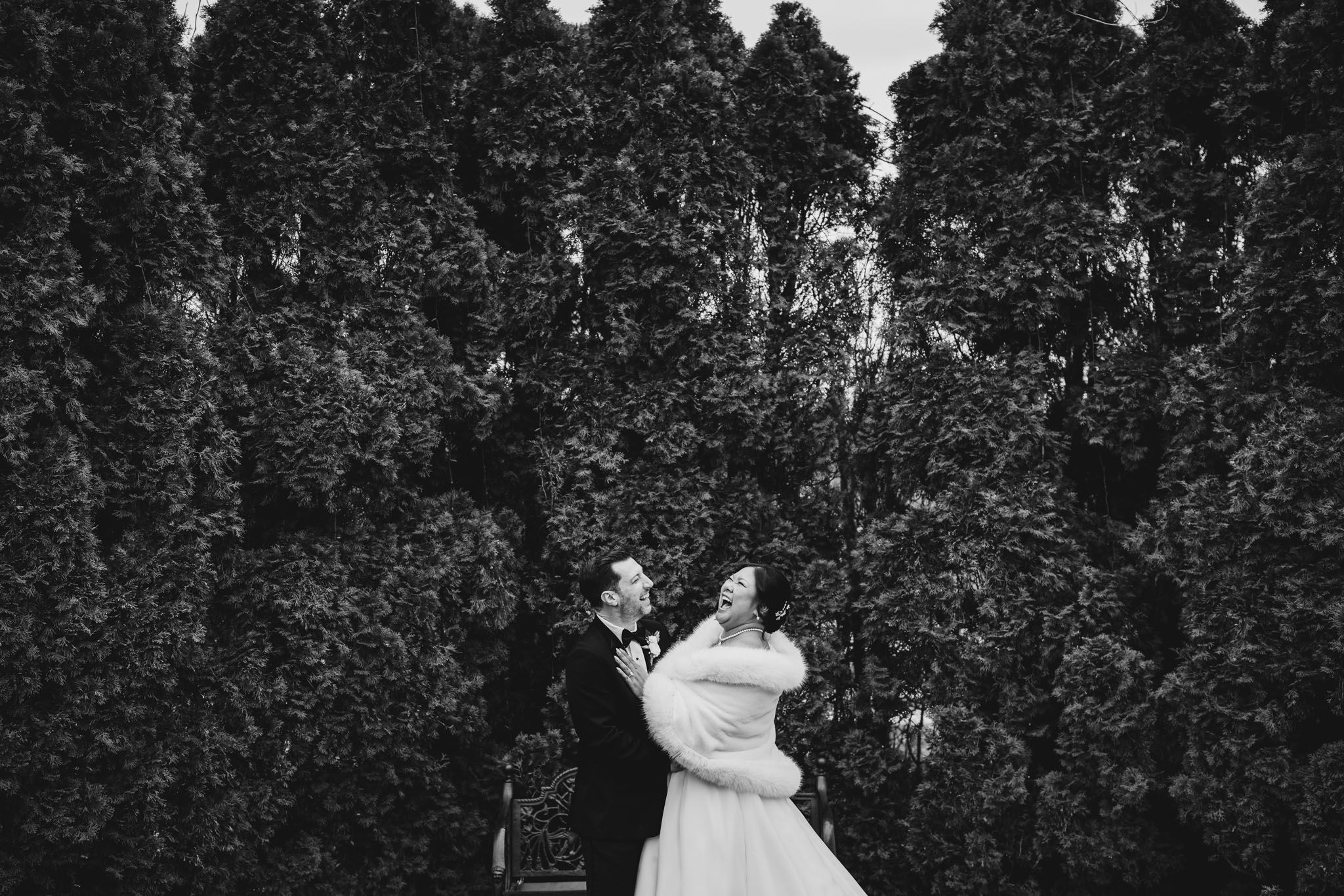 A romantic black and white wedding photo of a couple dancing among evergreen trees.