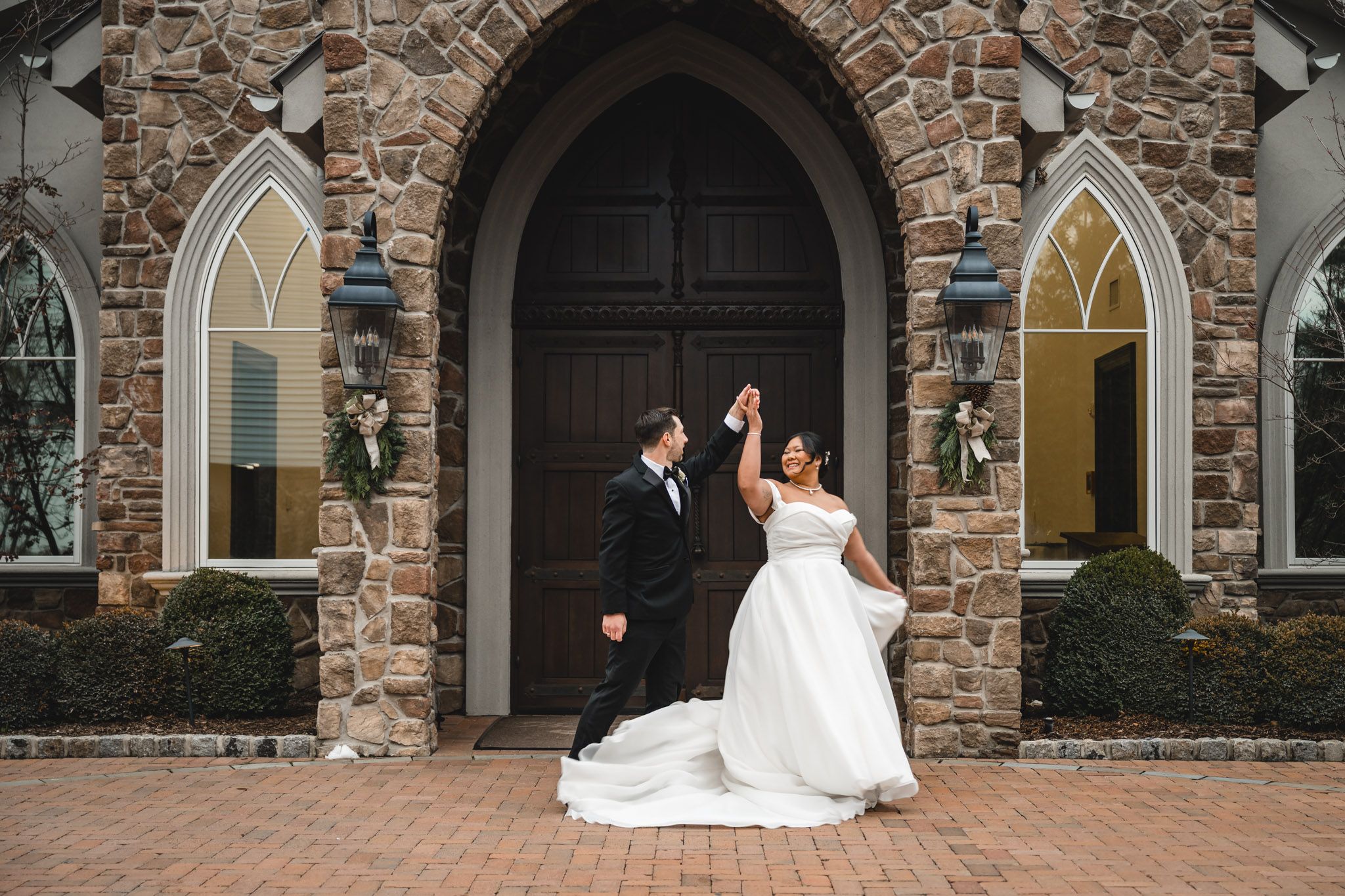 Wedding couple poses playfully in front of stone church with gothic arched doorway.