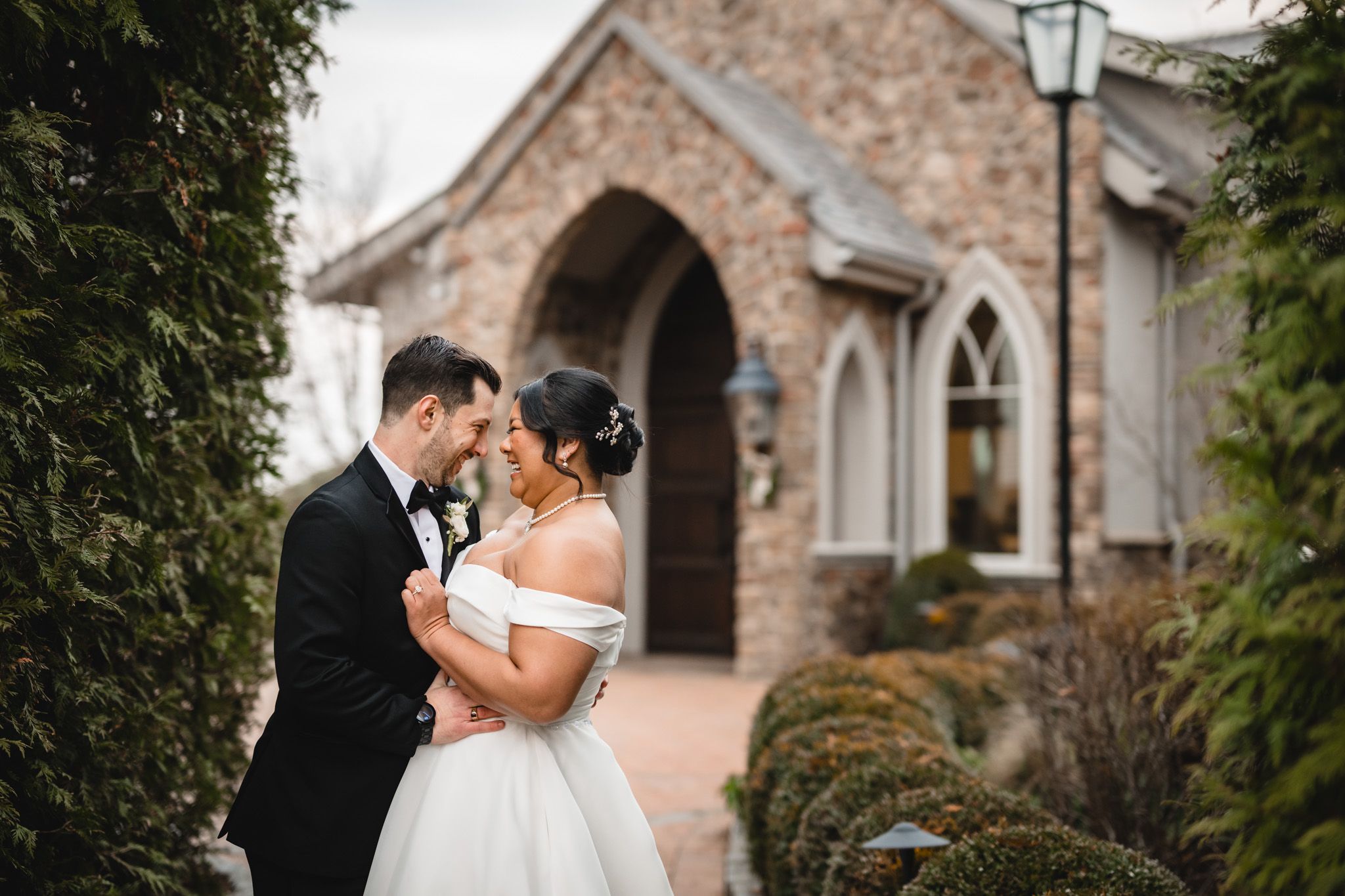 Elegant wedding portrait with stone chapel and evergreen landscaping in background.